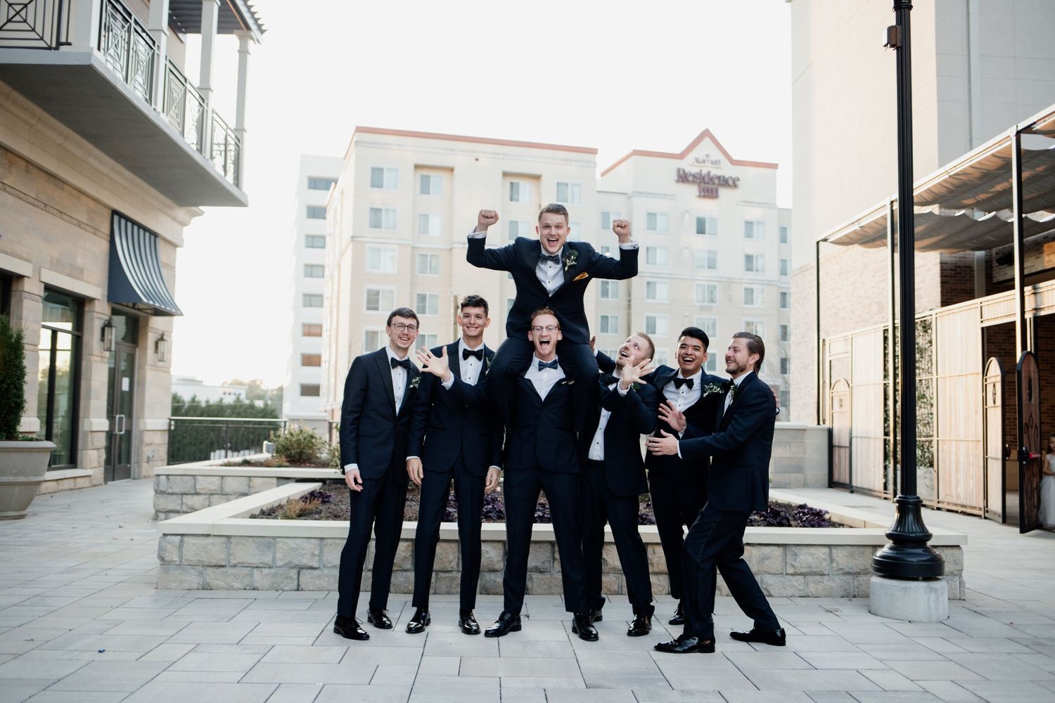 Wedding party in formal attire performs fun group lift pose outside urban building.