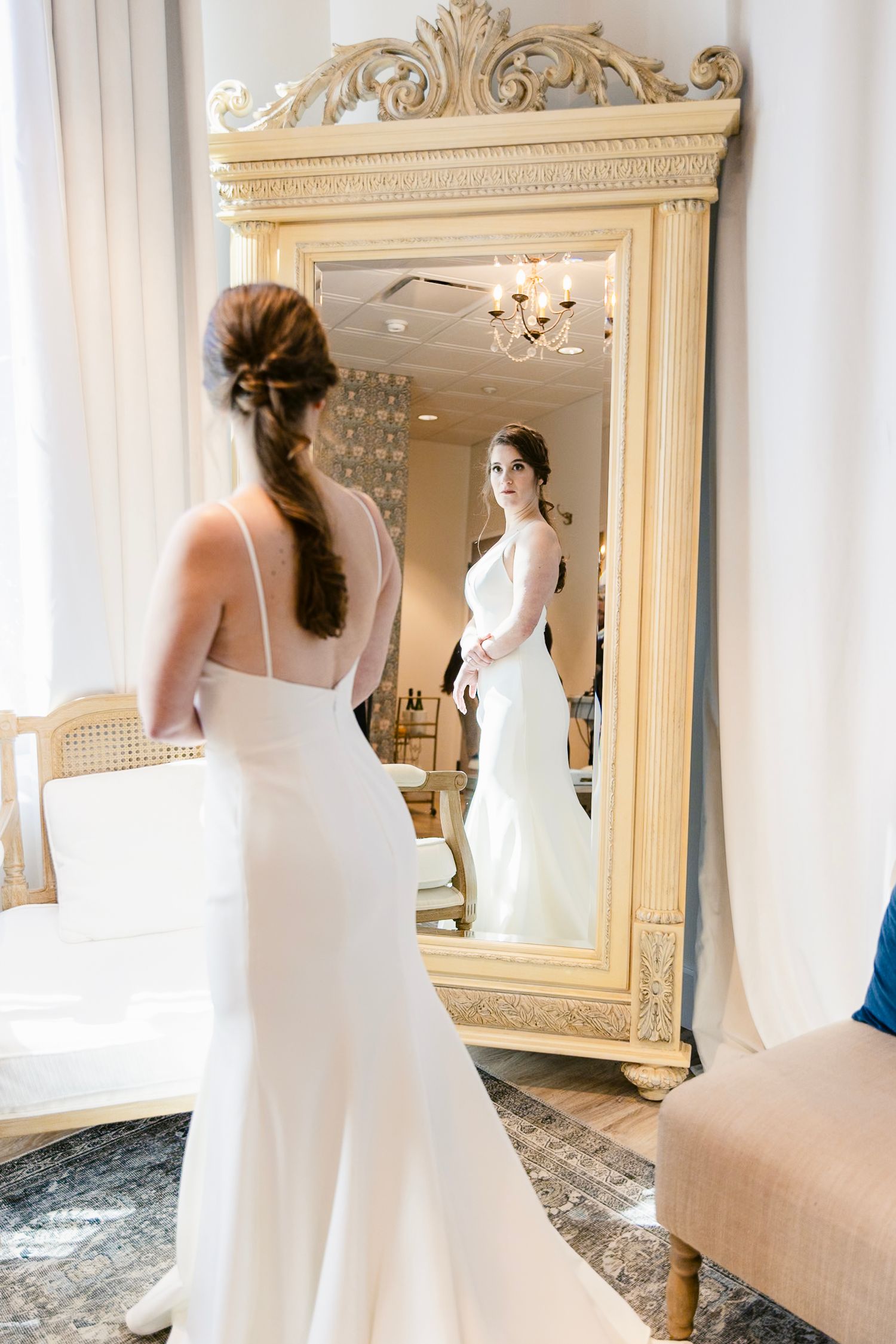 A bride in a white wedding gown looks at her reflection in an ornate cream-colored full length mirror.