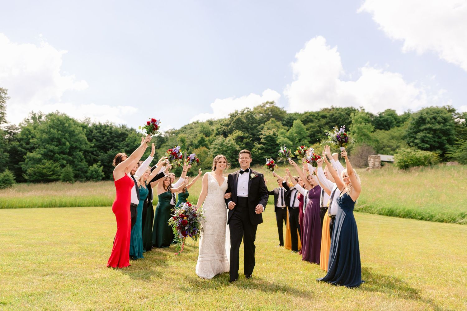 Joyful wedding party celebrating in sunny field with arms raised.
