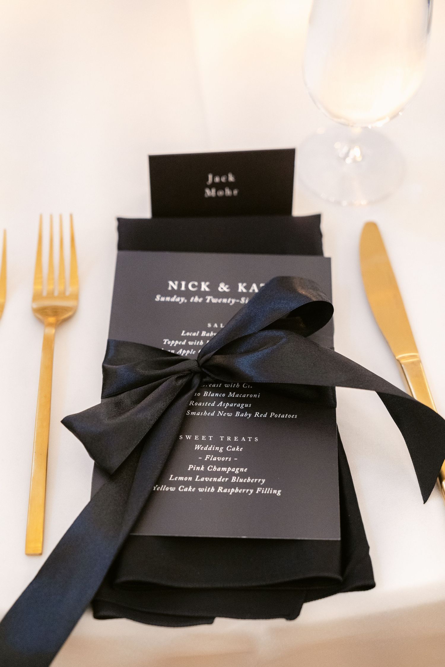 Black wedding menu with gold cutlery and dark bow tied ribbon set against white table setting.