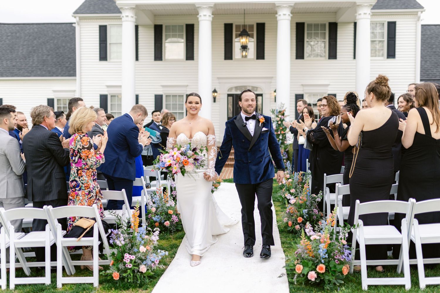 Wedding couple walking down flower-lined aisle outside white columned mansion.