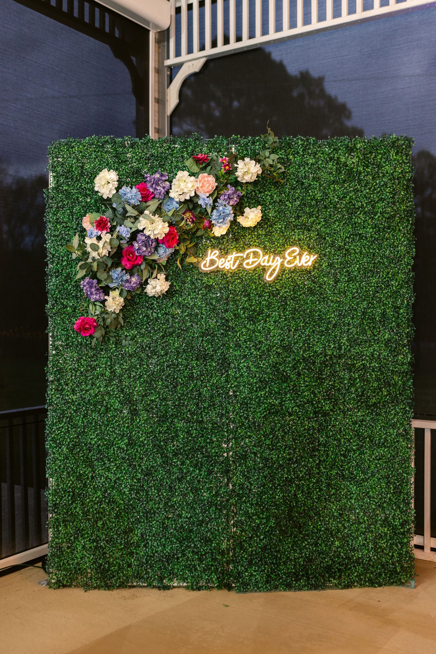 A green hedge wall backdrop with colorful flowers and neon 'Best Day Ever' sign for wedding photos.