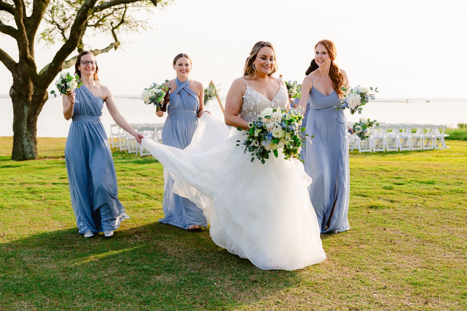Wedding party in light blue dresses walking together on a grassy field during sunset.