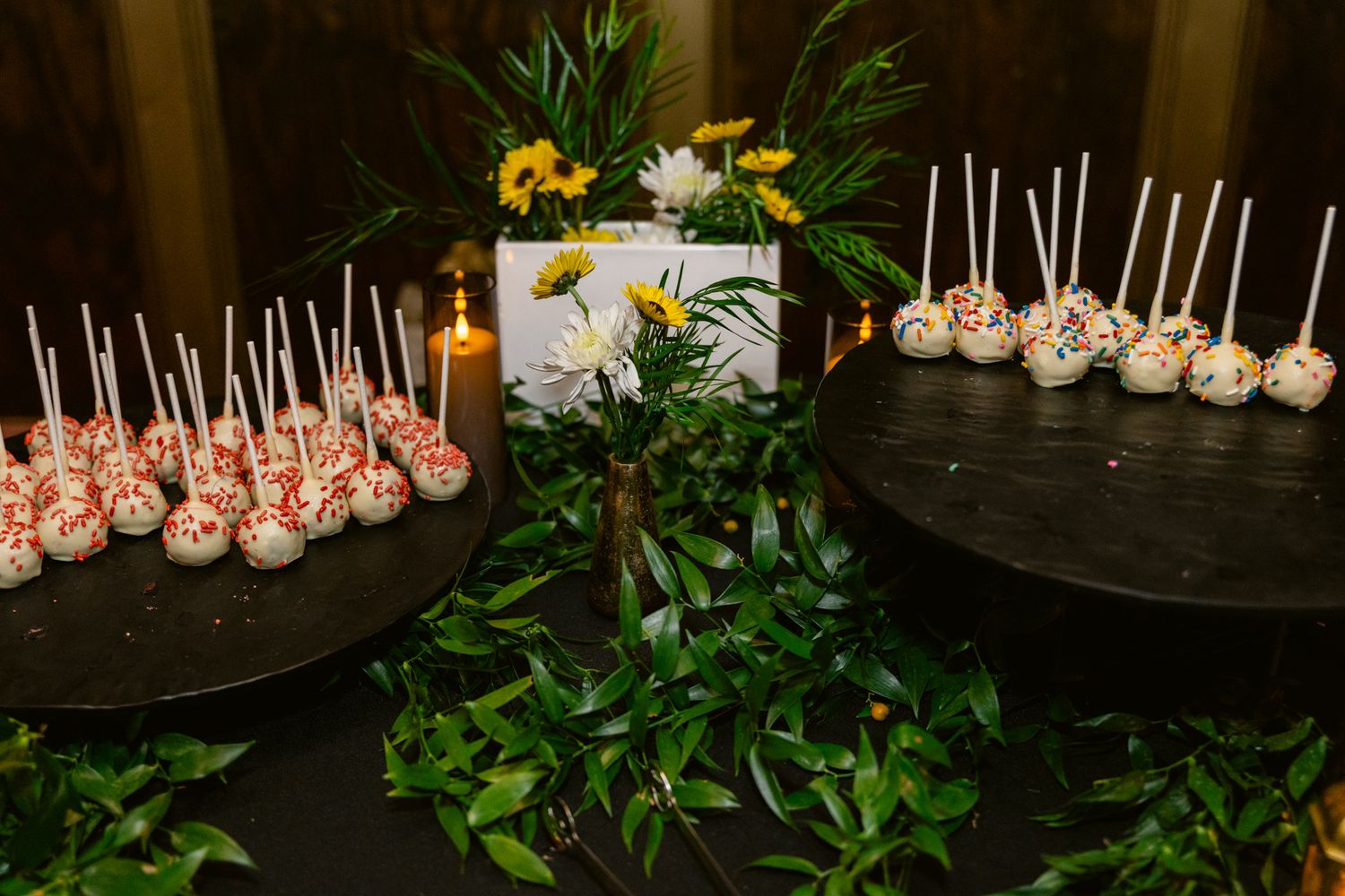 Pink and white cake pops displayed on a dark table with green foliage decorations in the background.