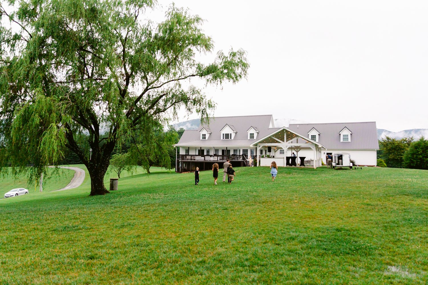 A large white barn-style building with a wraparound porch sits on a lush green lawn with a weeping willow tree.