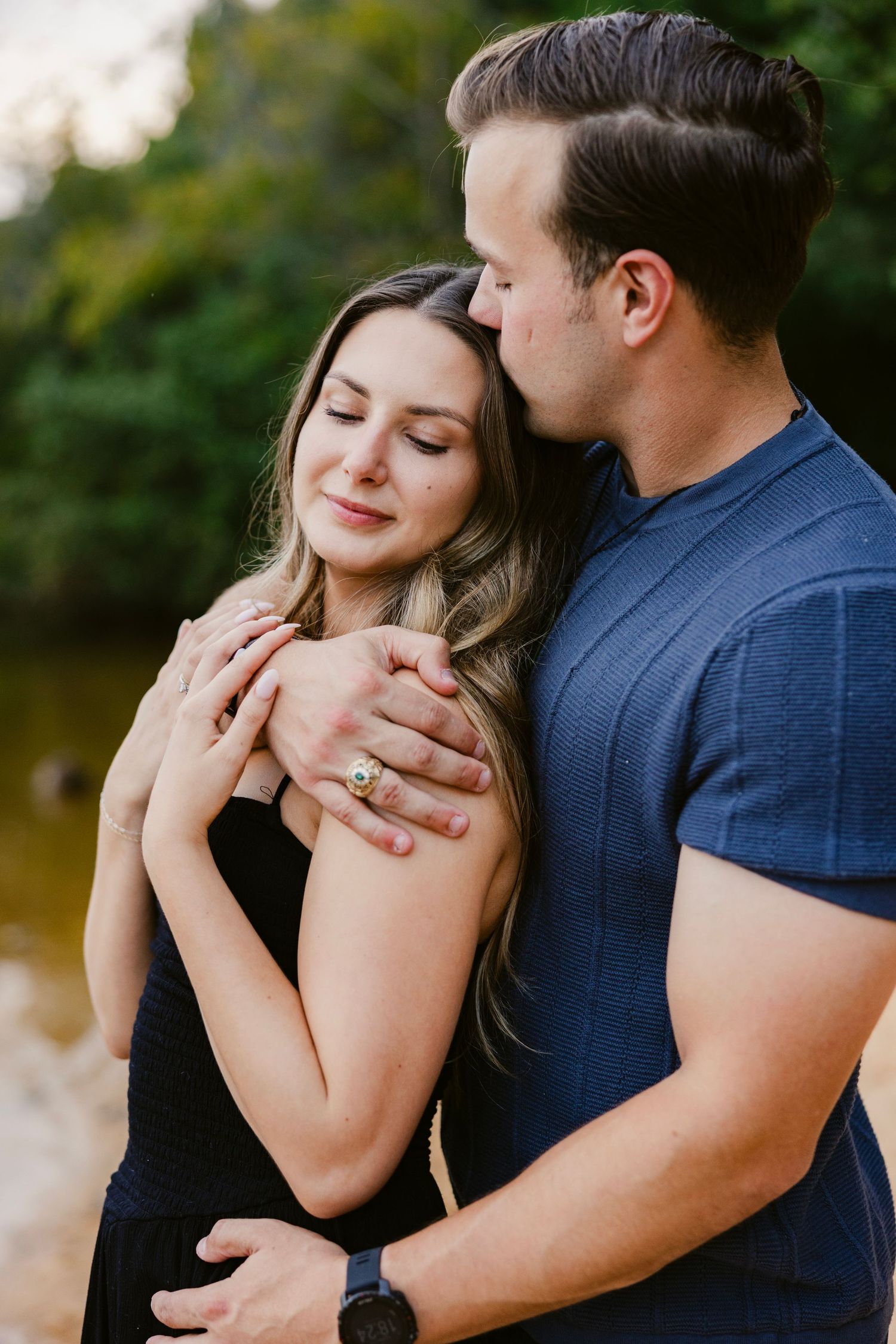 A romantic couple embraces outdoors, with her eyes closed and wearing a black dress while he wears a navy blue shirt.