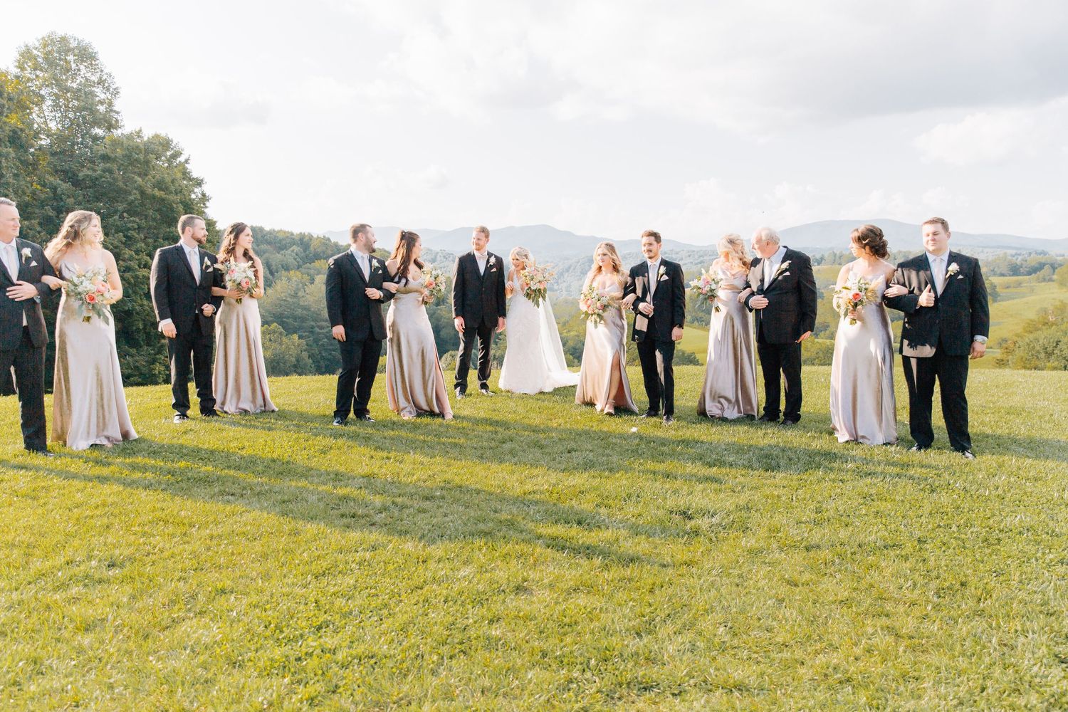 Wedding guests gather on a grassy hilltop during a scenic outdoor ceremony in warm sunlight.