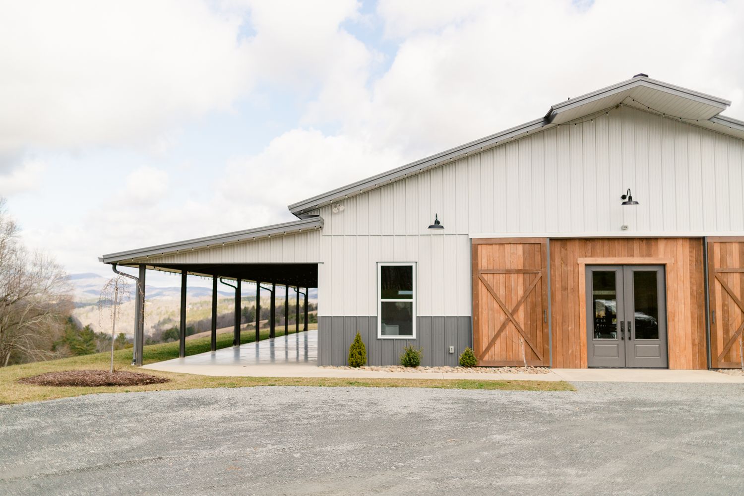 A modern white barn-style building with wooden accents and a long covered porch area extends across a rural landscape.