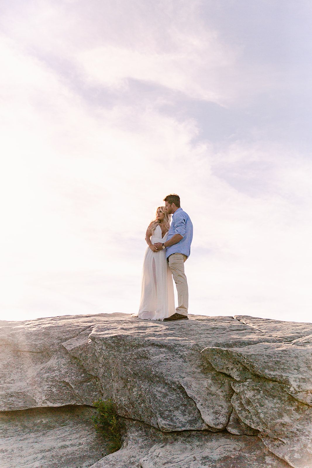A couple in wedding attire shares an intimate moment while standing on a large rocky outcrop against a cloudy sky.