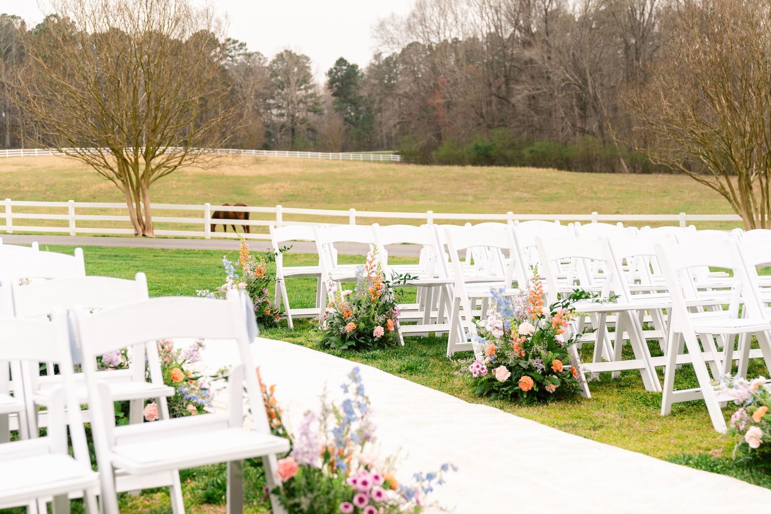 White wedding ceremony chairs arranged in rows with colorful floral arrangements along the aisle in an outdoor garden setting.