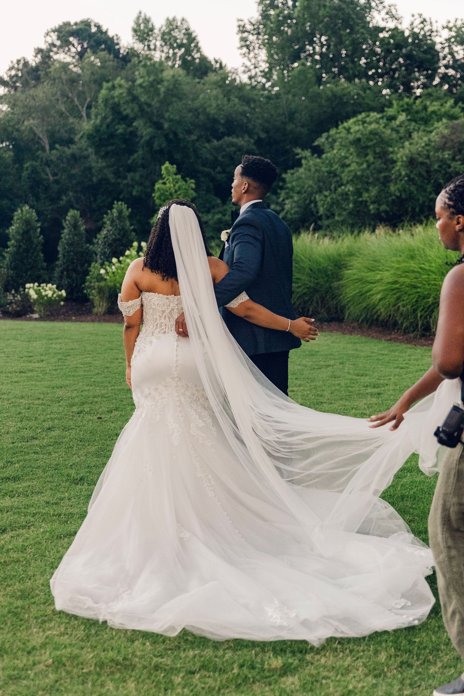 A bride in a flowing white wedding dress with a long veil walks on lush green grass during an outdoor ceremony.