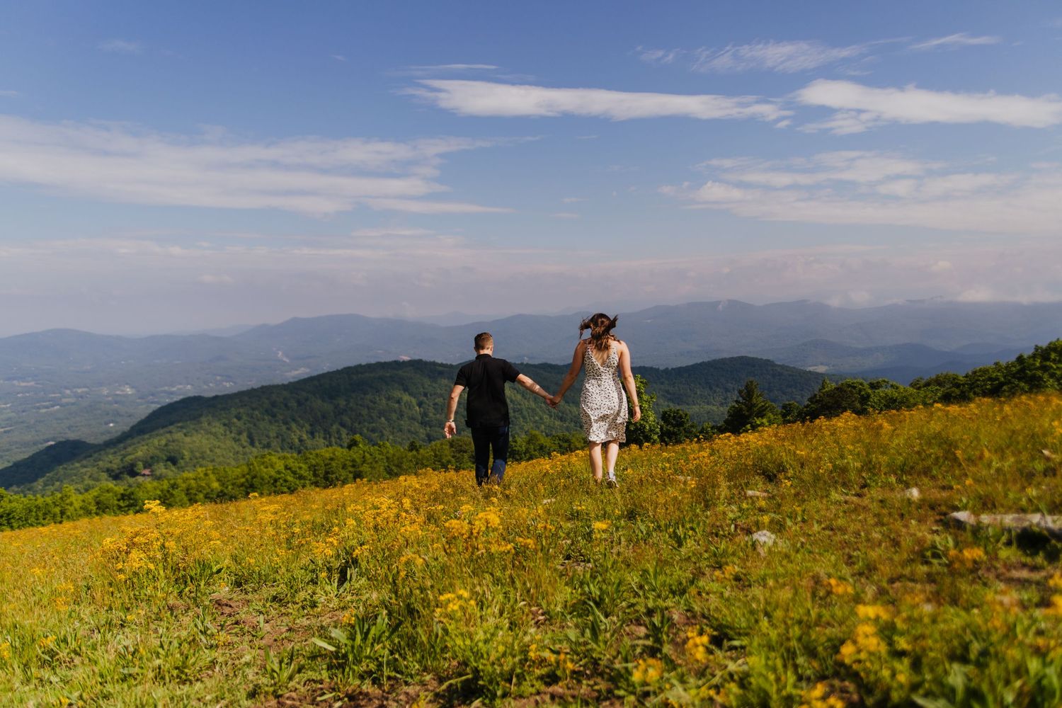 Couple walks hand in hand through a wildflower meadow on a mountaintop with scenic valley views.