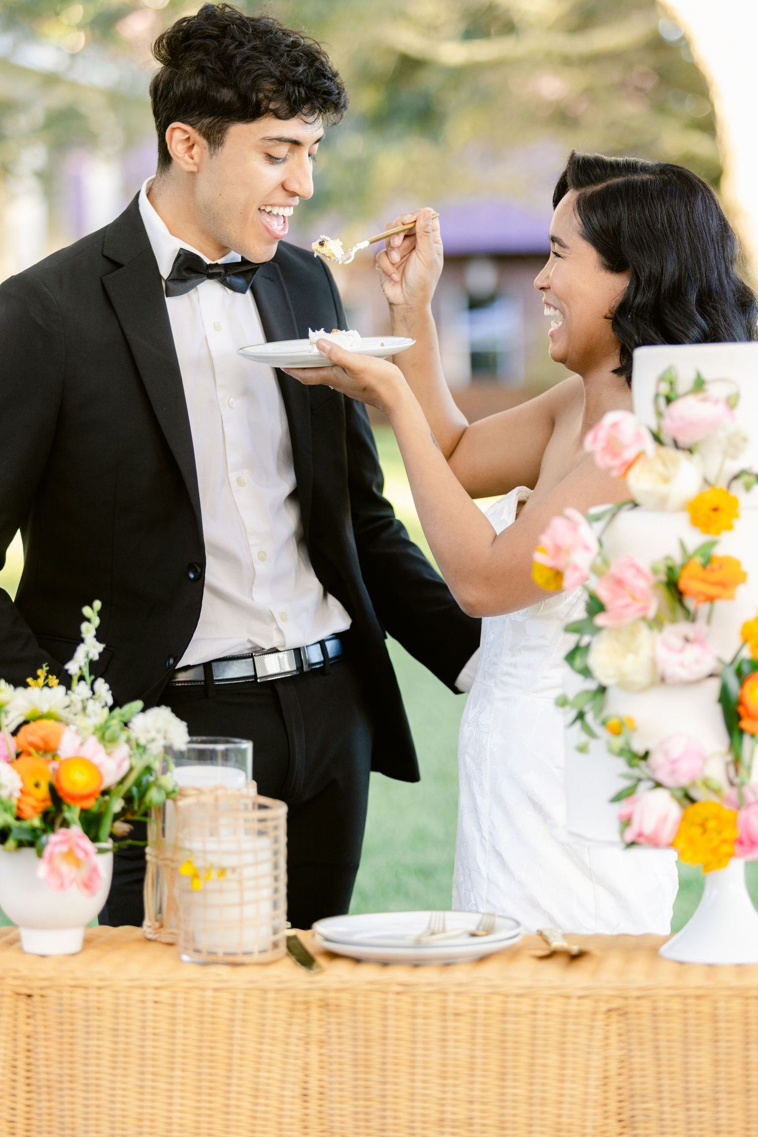 Bride feeds groom wedding cake at outdoor reception table decorated with colorful pink and orange floral arrangements.