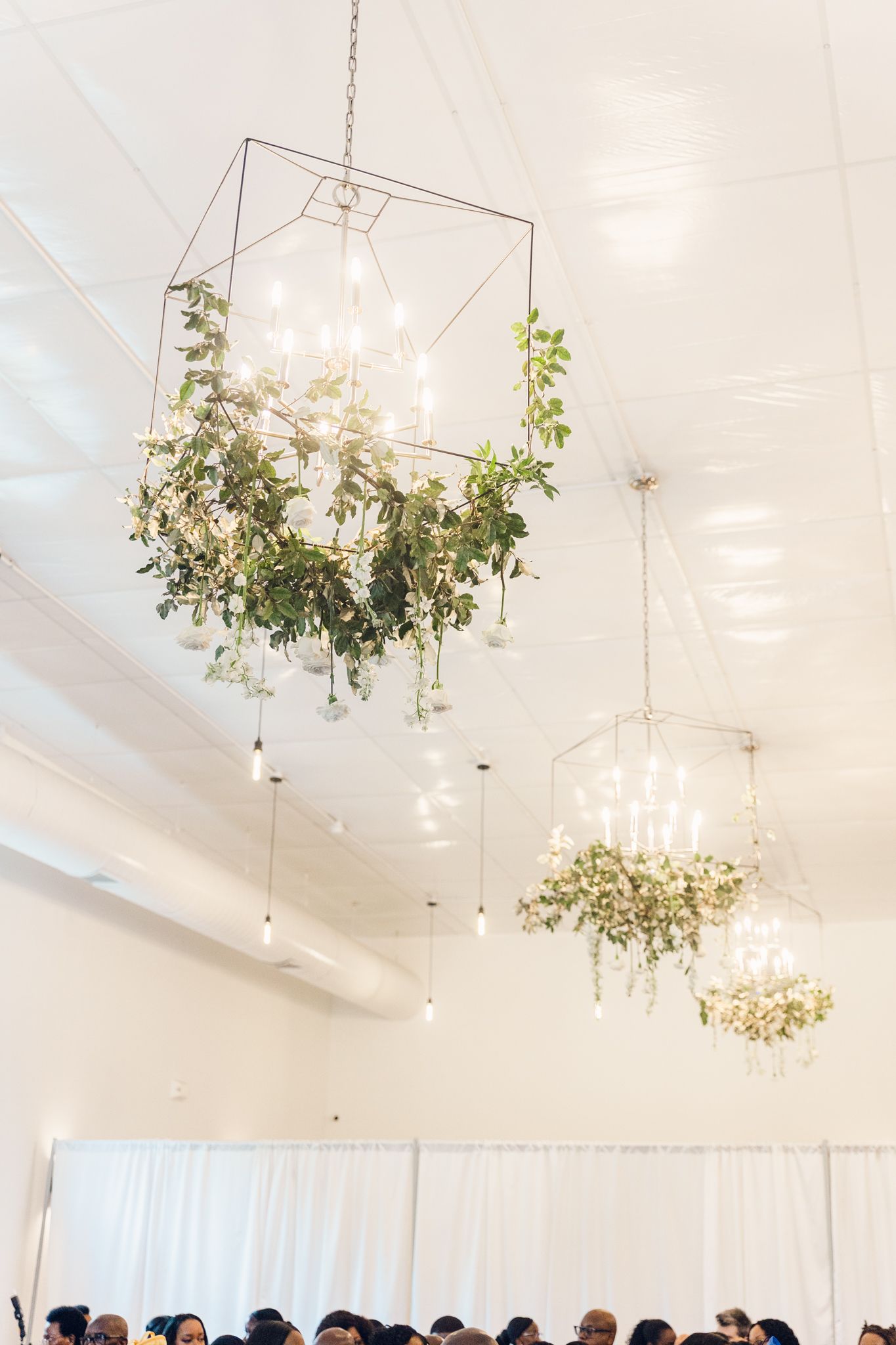 Modern geometric chandeliers adorned with cascading greenery hanging from a white ceiling in an event space.