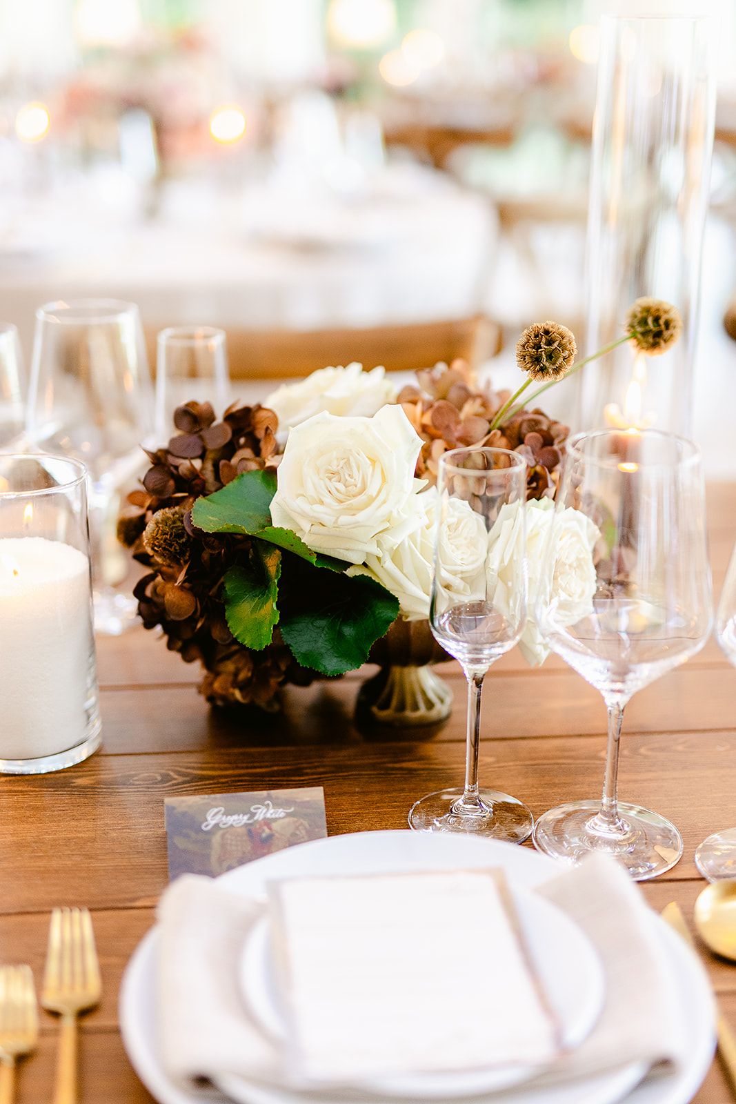 Close-up of formal place setting with white roses, wine glasses, and elegant tableware on wooden surface.