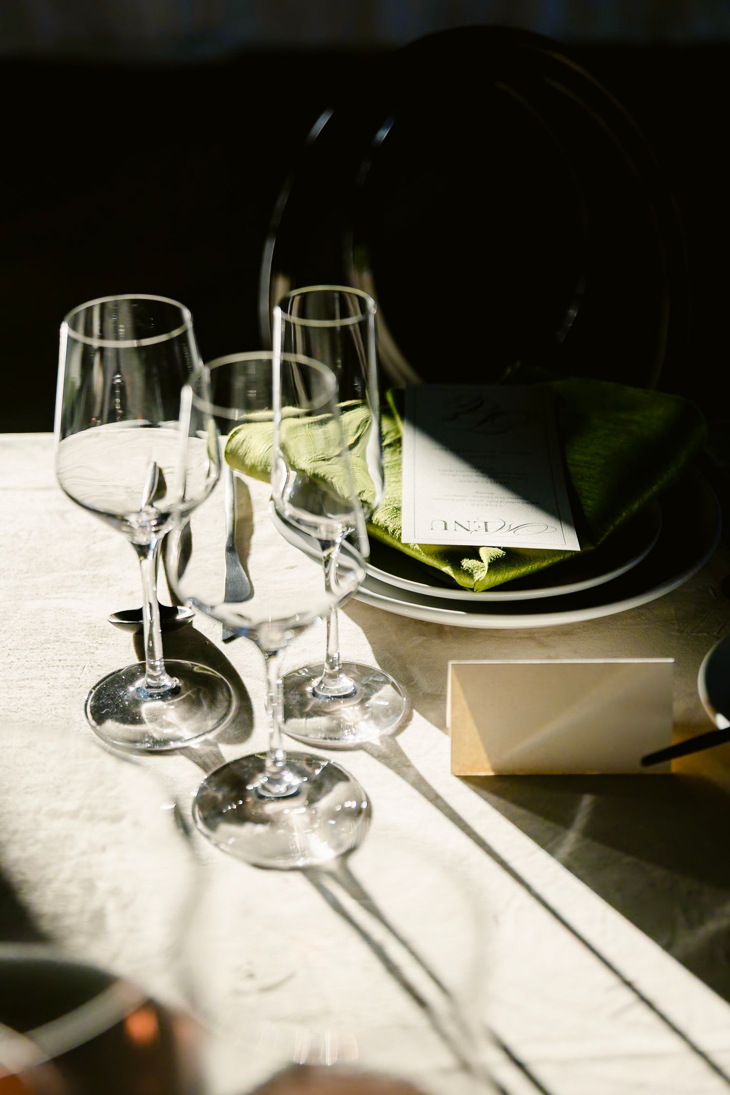 Wine glasses casting dramatic shadows on a white tablecloth in natural sunlight.