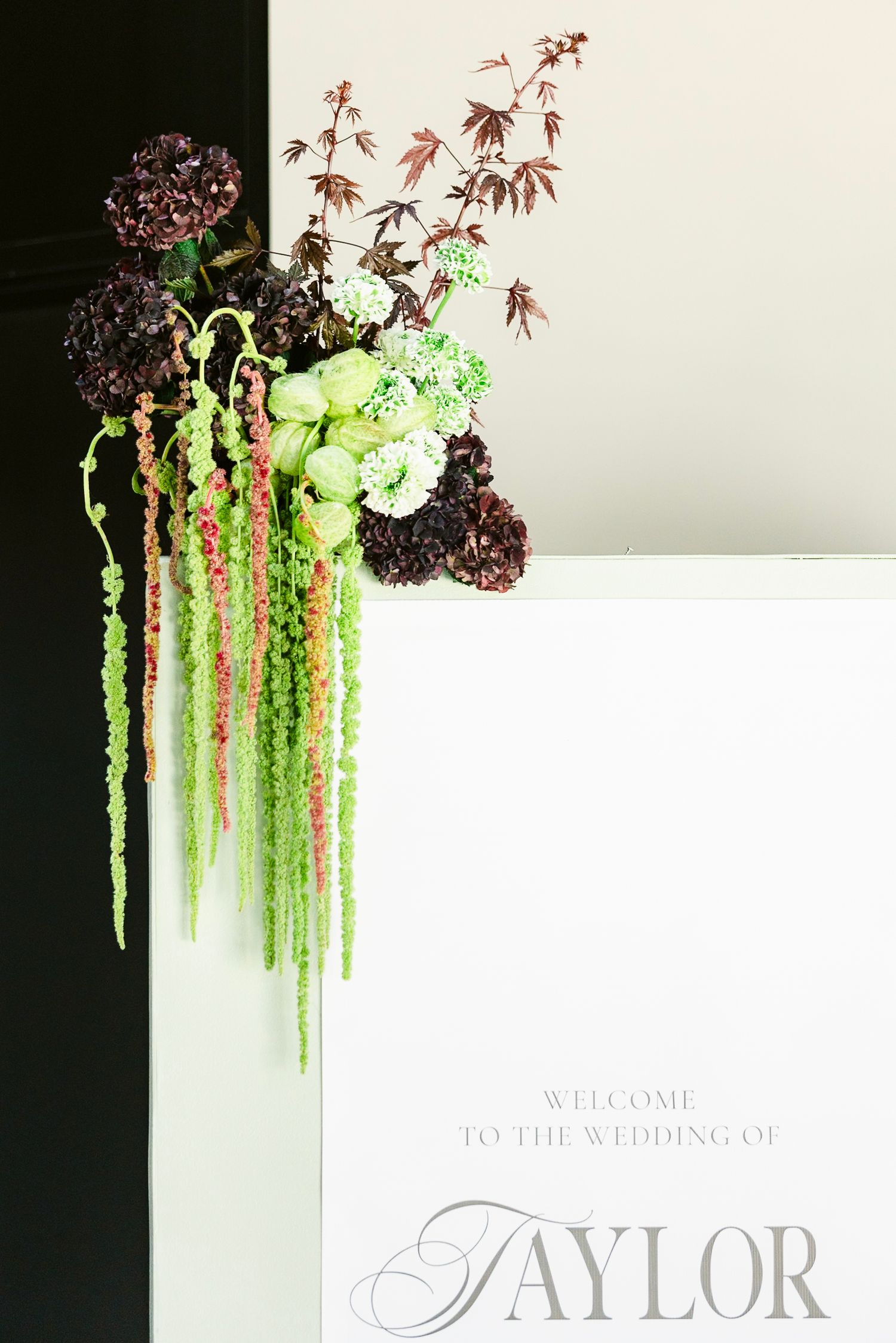 Elegant floral arrangement with cascading green amaranthus and burgundy blooms adorns a wedding welcome sign.
