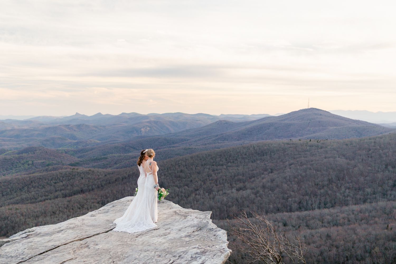 A couple in wedding attire stands on a mountain summit overlooking a scenic mountain range at sunset.