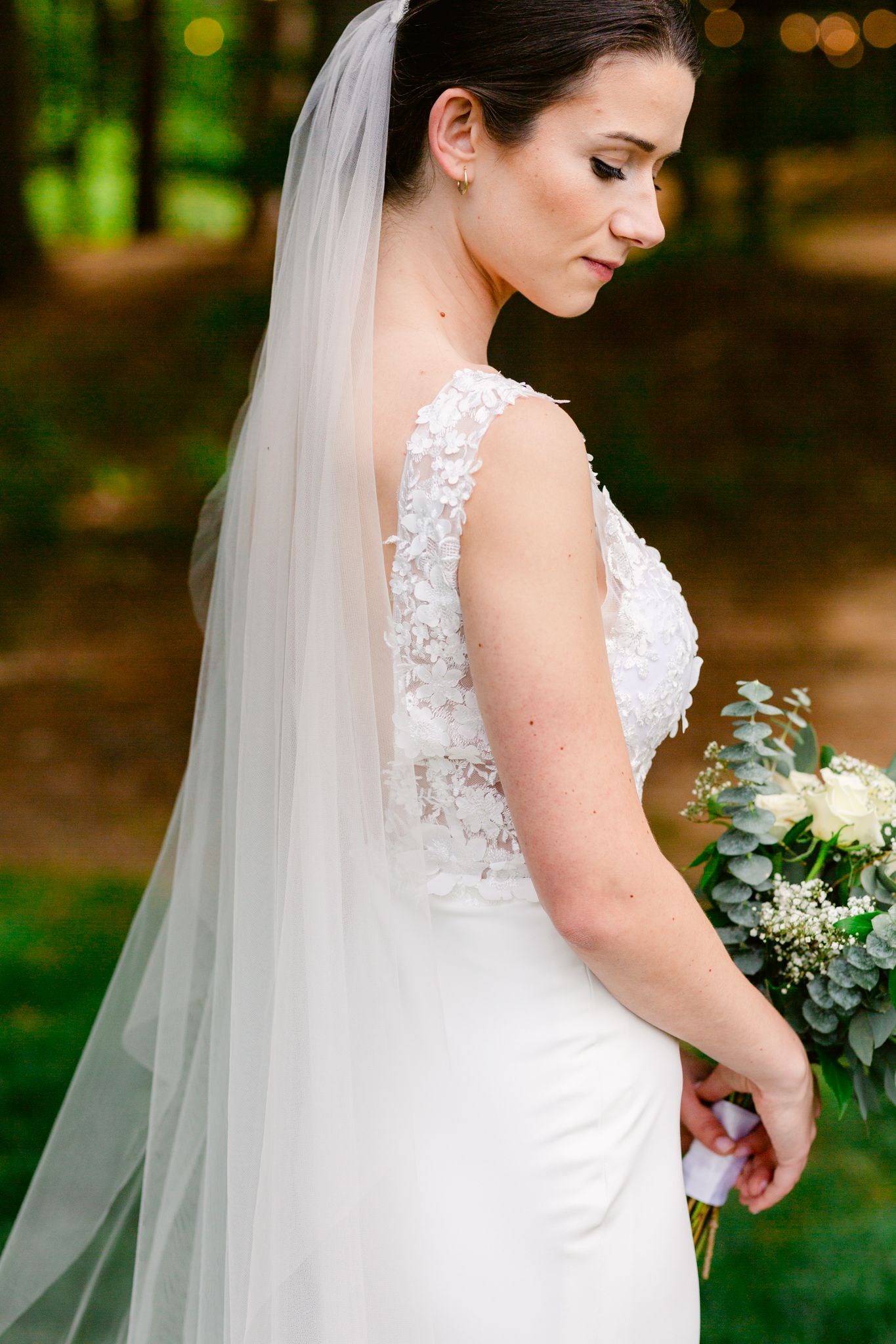 Bride in elegant lace wedding dress with flowing veil poses in a garden setting with white floral bouquet.