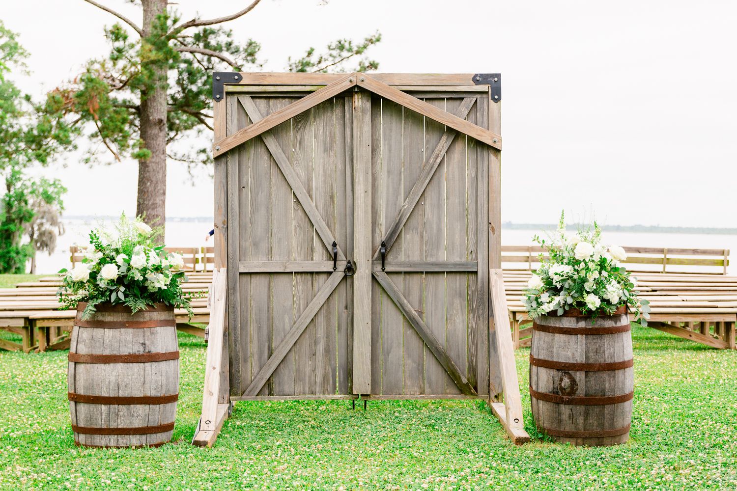 Rustic wooden barrels and barn door decor create a charming outdoor wedding setting in a grassy field.