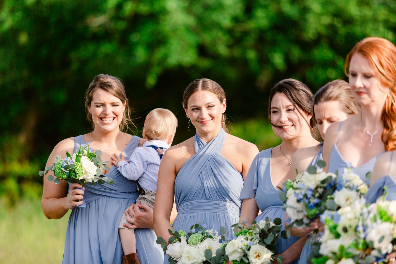 Wedding party in light blue dresses poses outdoors with floral bouquets in a garden setting.