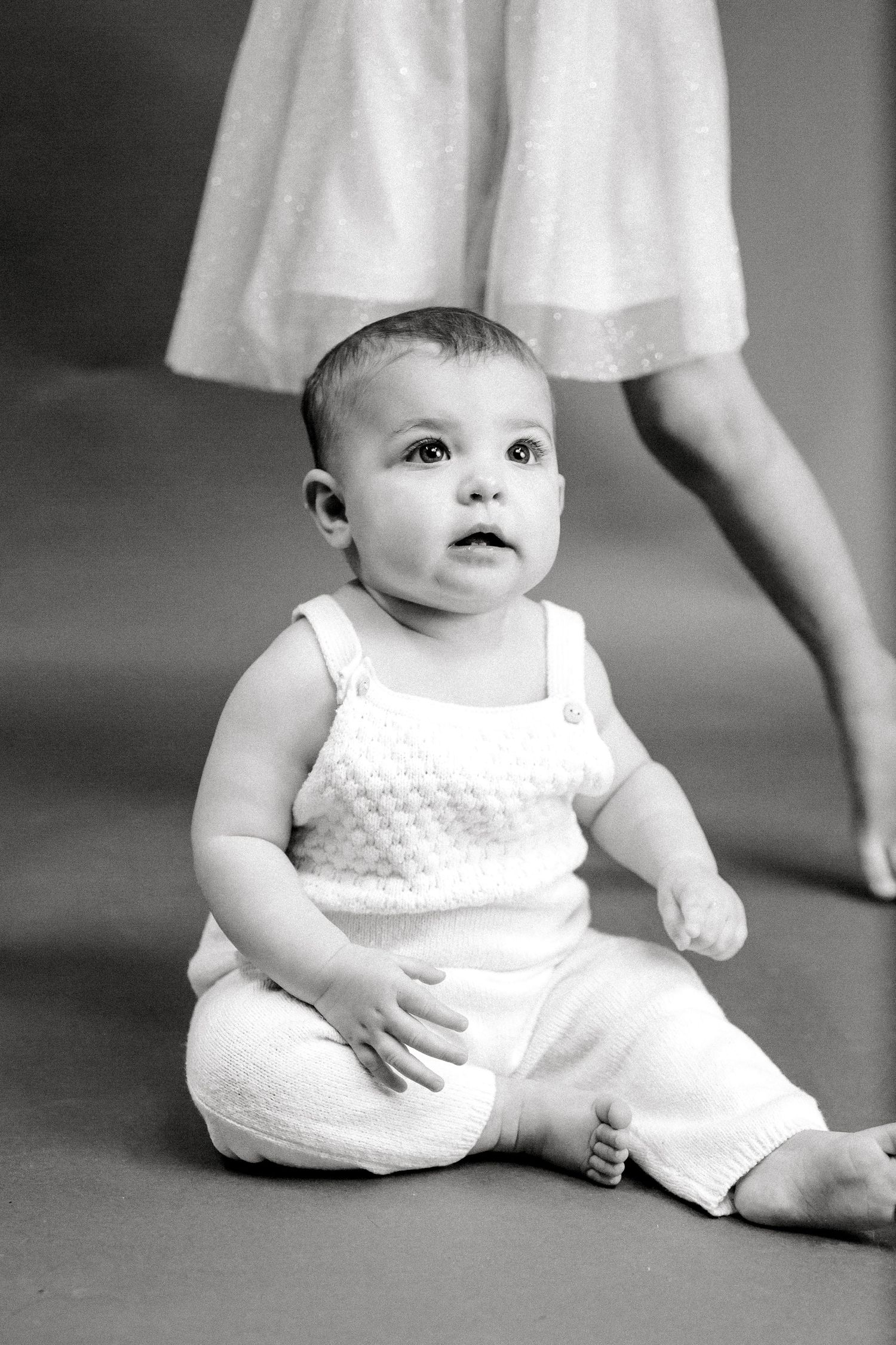 Baby in white knit romper sitting on gray floor with adult feet visible.