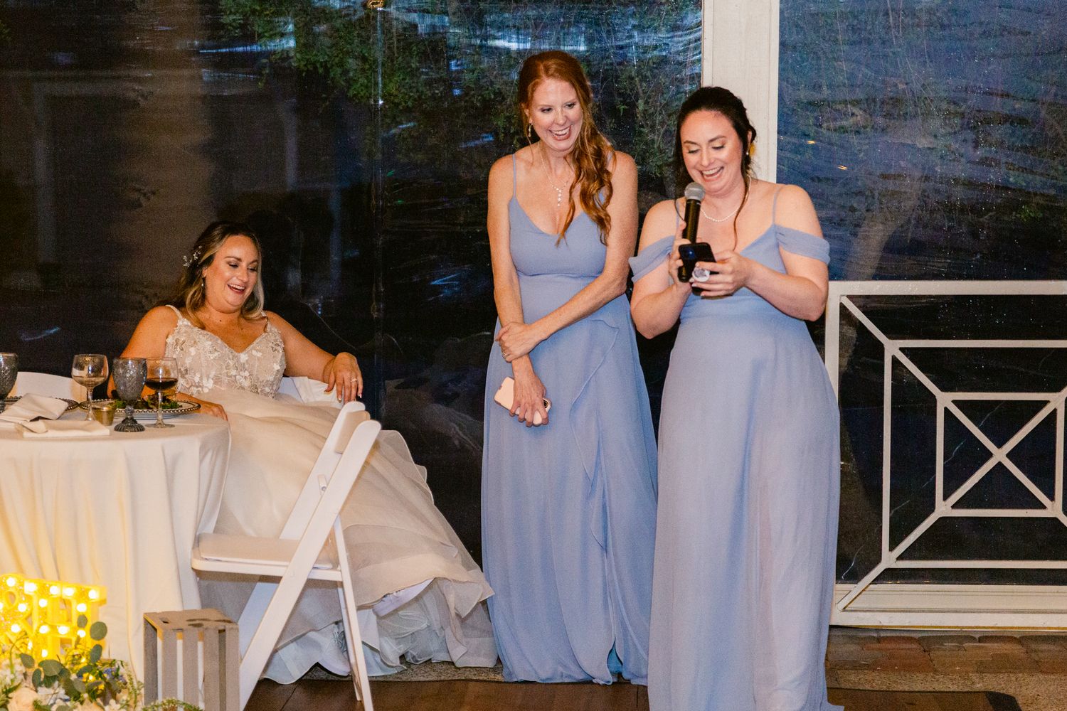 Wedding party in light blue dresses poses together at an outdoor venue with stone walls and floral decor.
