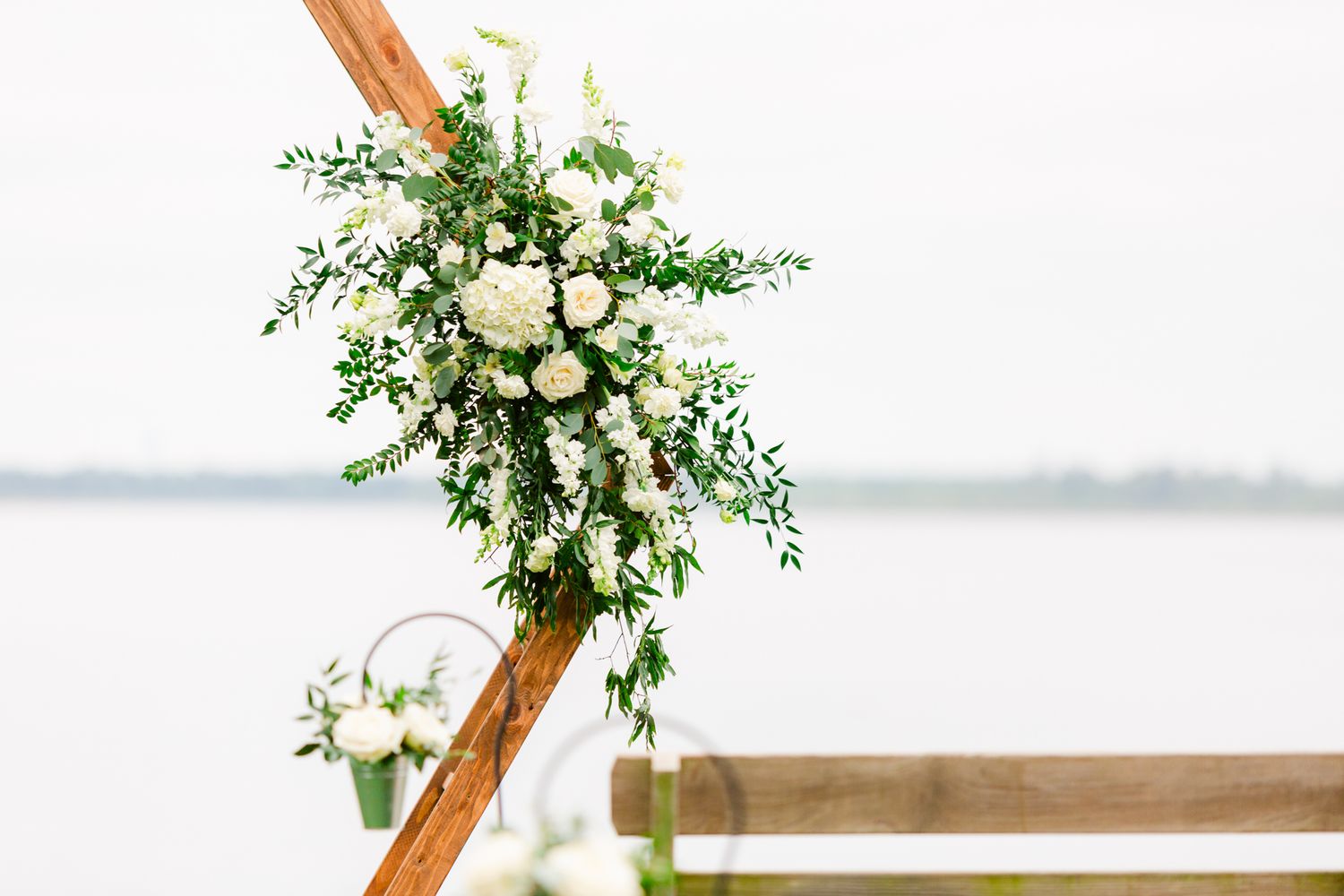 A rustic wooden arch decorated with white roses and greenery stands against a waterfront backdrop.