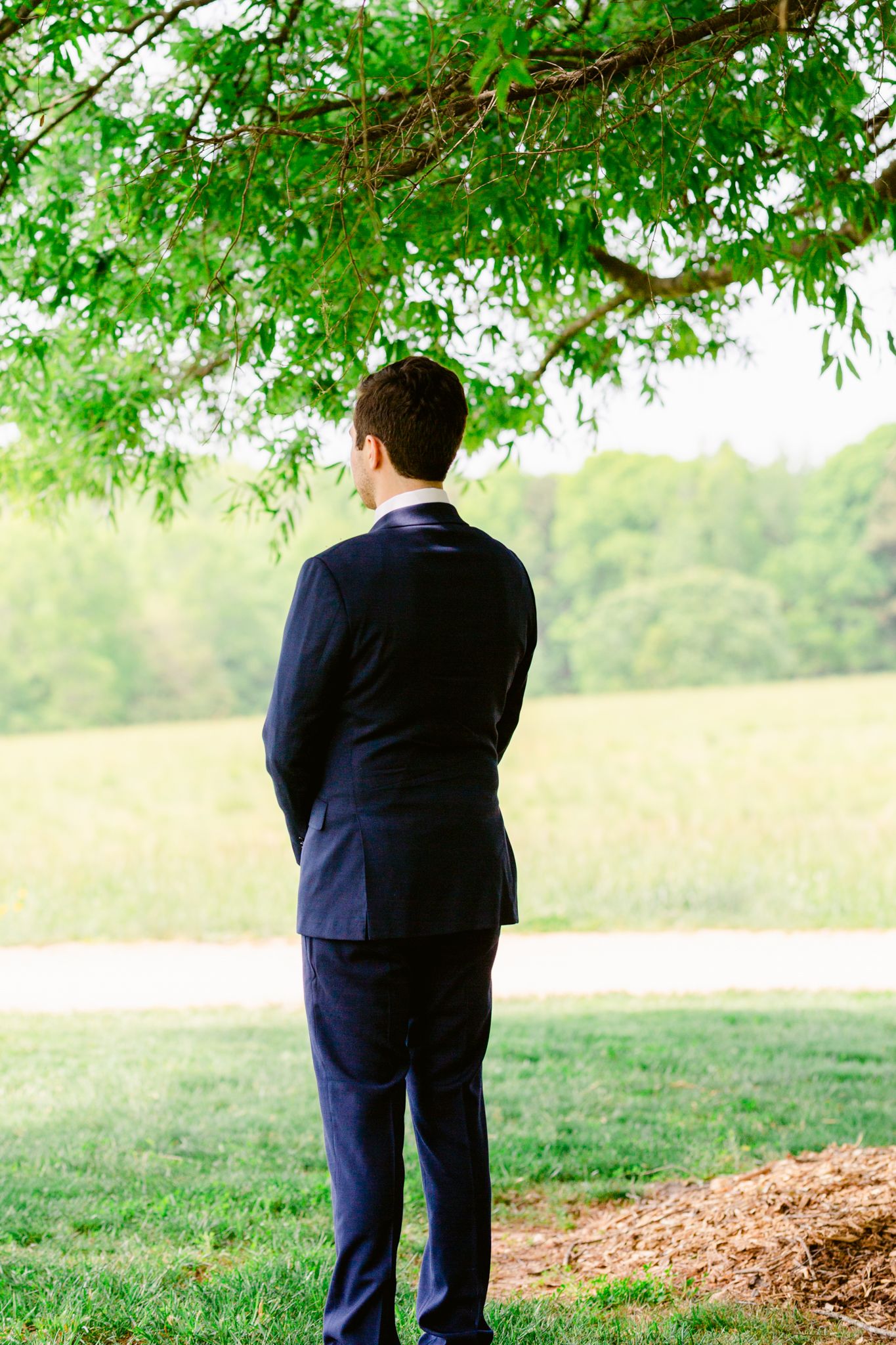 A person in a navy suit stands near a dirt mound under a green leafy tree in a pastoral outdoor setting.