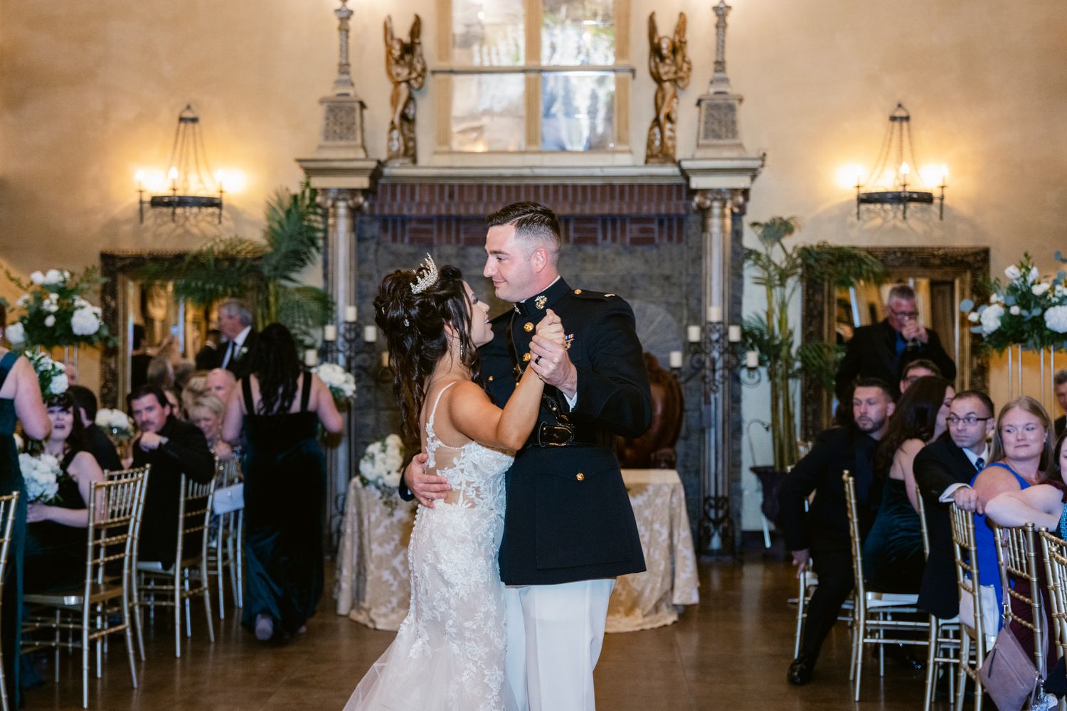 Couple dancing together at an elegant wedding reception in a grand ballroom with chandeliers and guests watching.