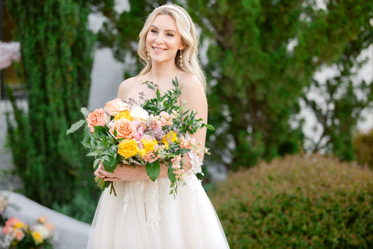 A bride in a white wedding dress holds a colorful bouquet of peach and yellow flowers with greenery in a garden setting.