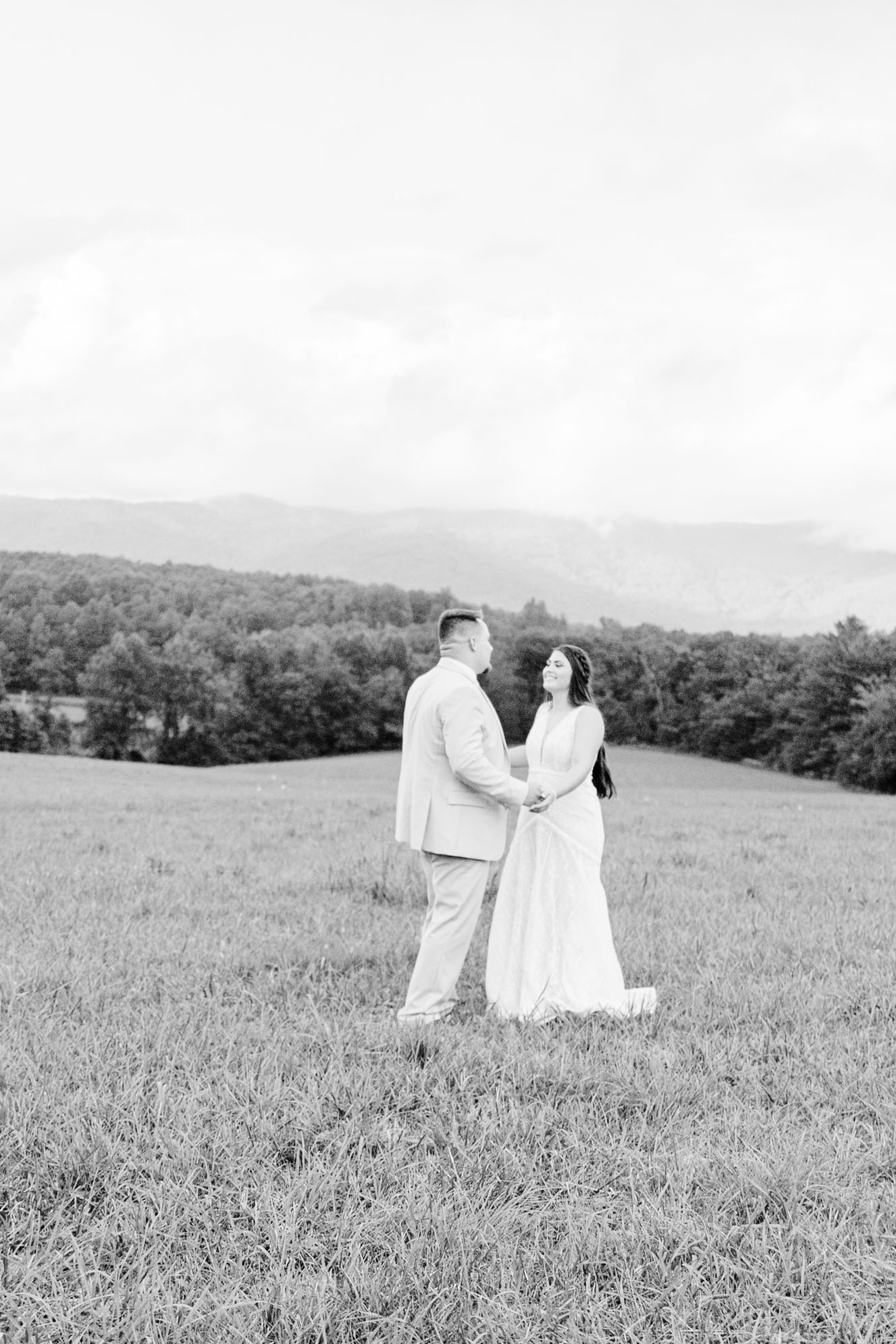 A black and white photograph captures a couple walking through a serene hillside meadow.