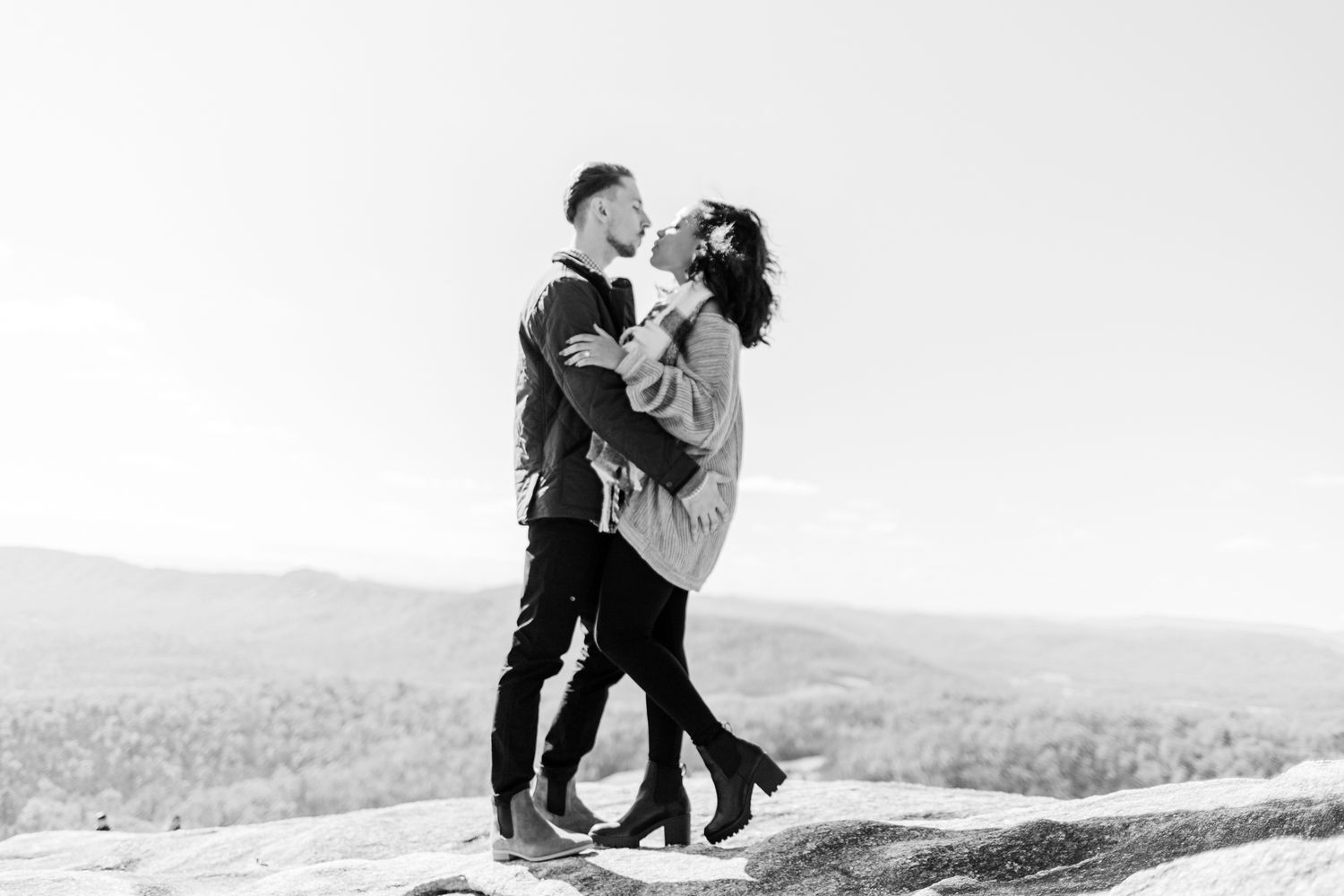A black and white photo captures a couple sharing an intimate moment on a mountain ledge.