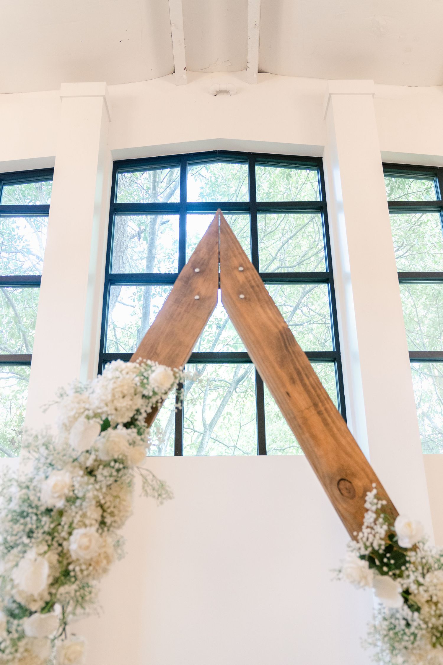 Wooden arch decorated with white floral arrangements in a modern white venue with large windows.