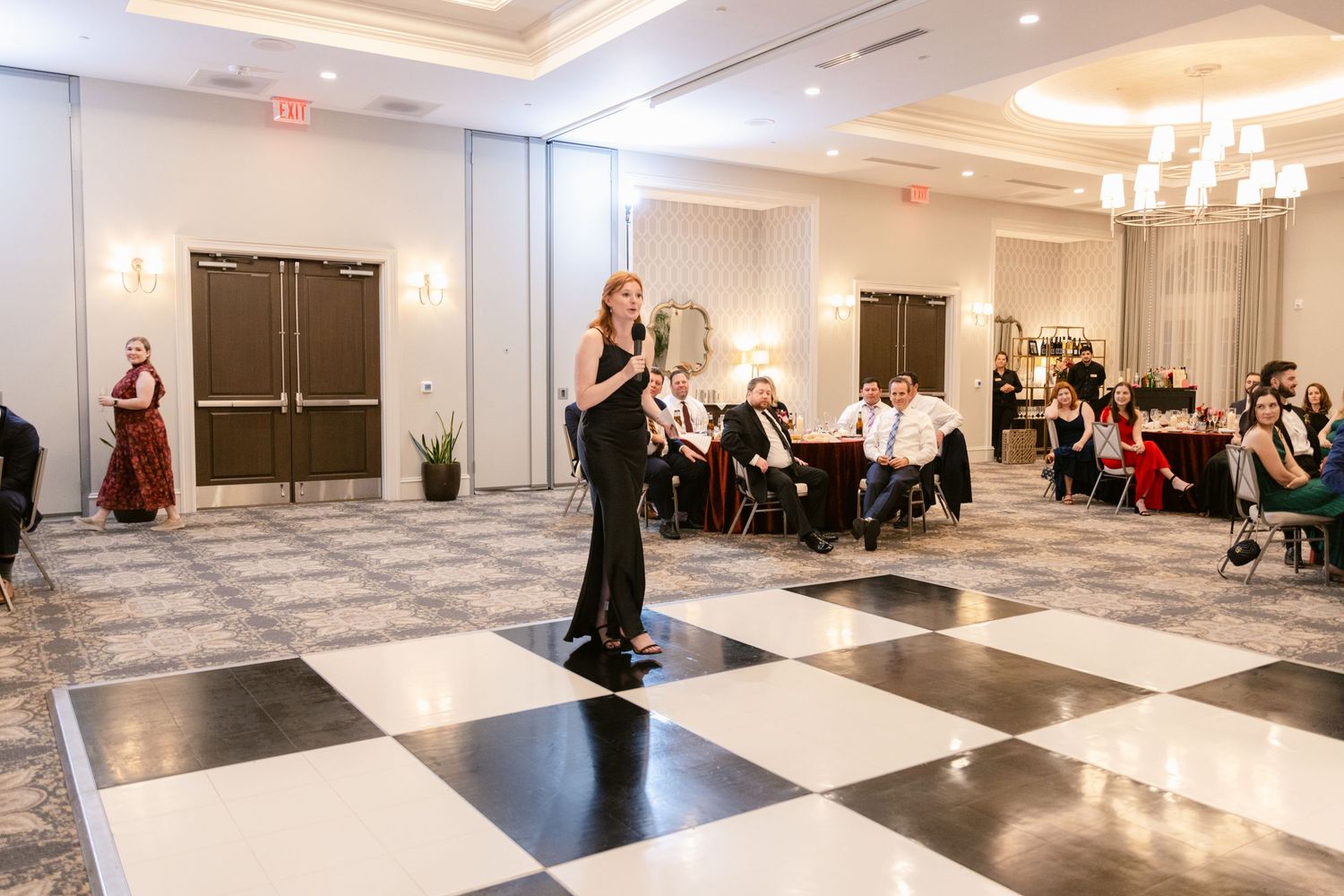 Guests watch and capture moments during a wedding reception in a modern white ballroom with checkerboard flooring.