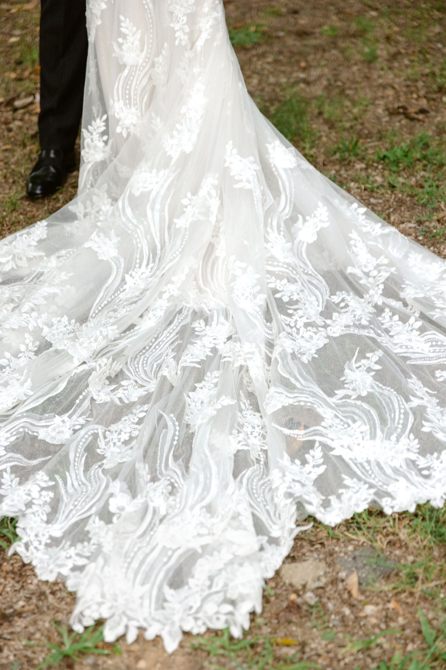 Close-up details of an elegant white lace wedding dress train spread across natural outdoor ground.