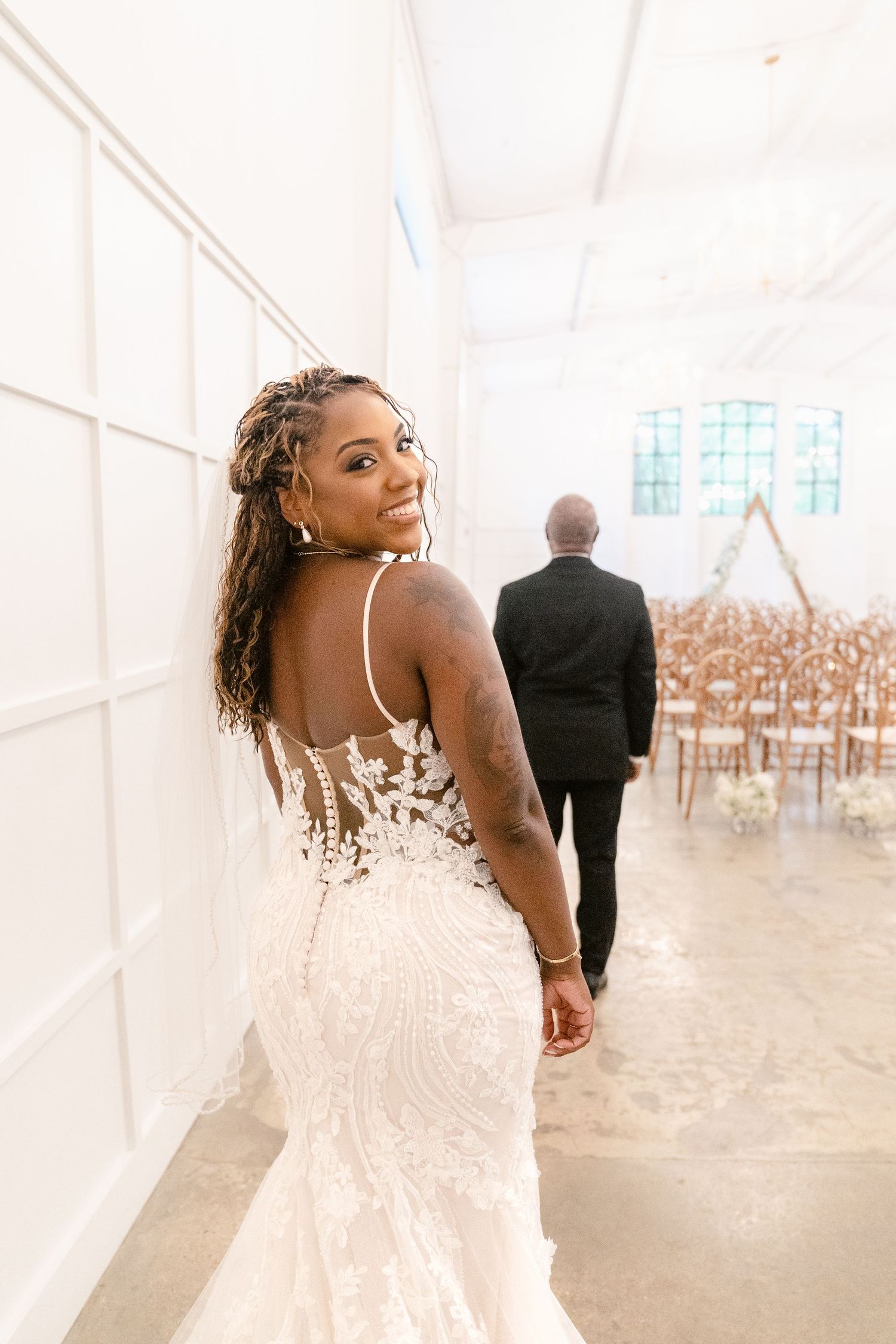 A bride in a white lace wedding dress poses gracefully in a bright, airy reception tent with rows of chairs.