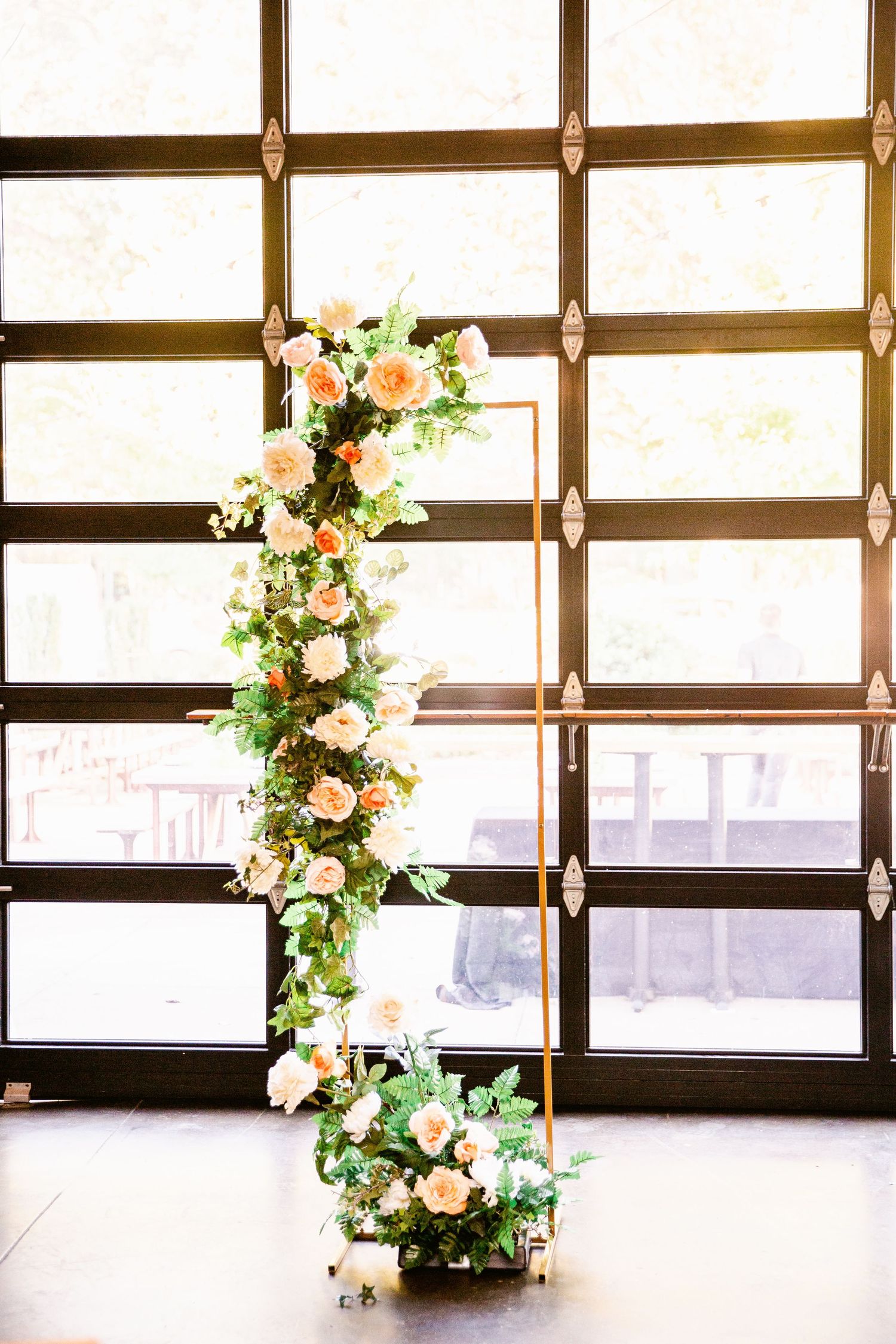 A curved floral arrangement with peach roses and greenery stands against a geometric window grid in natural light.
