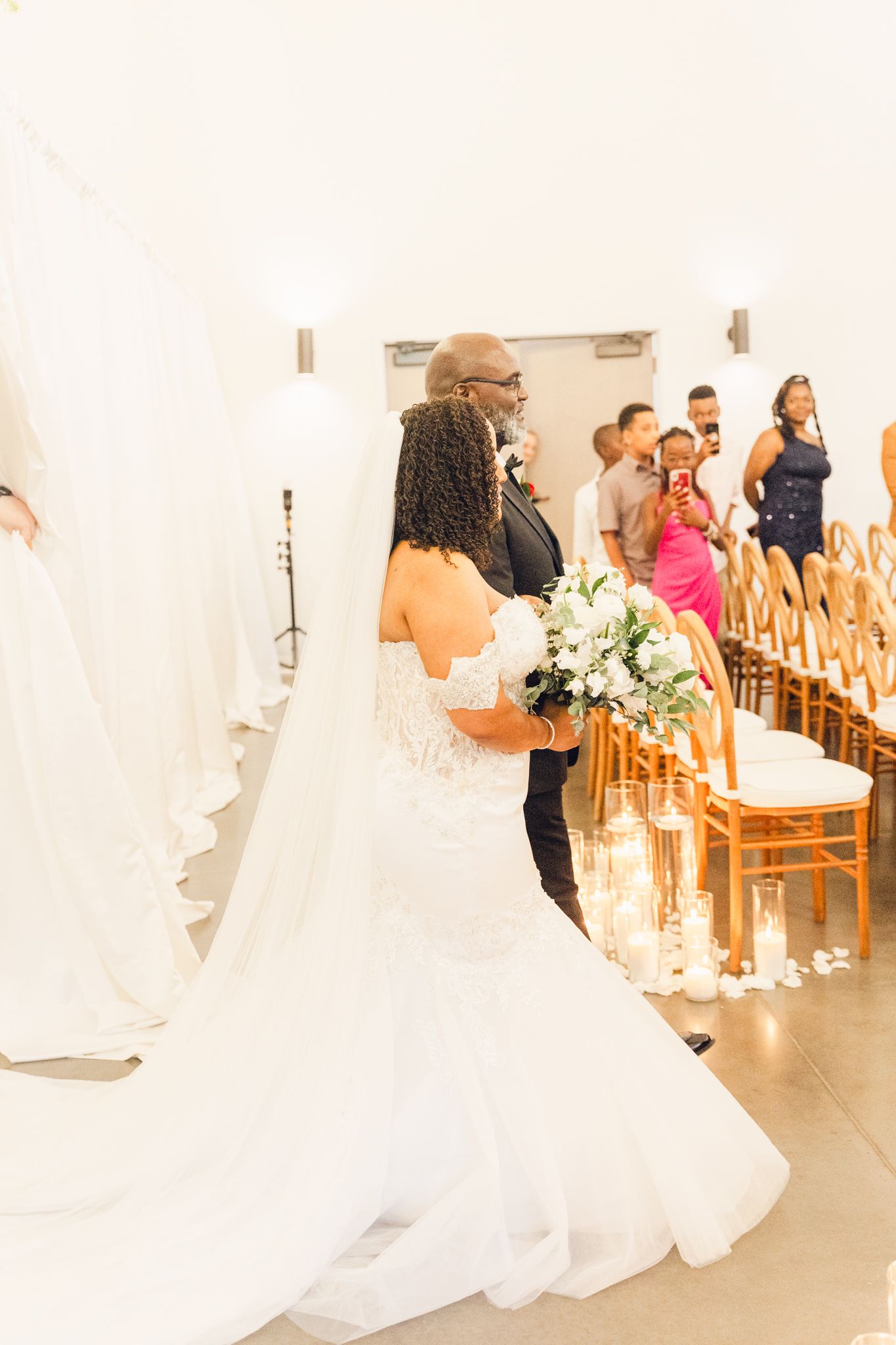 Wedding guests gather in an elegant venue with wooden chairs and white decor for the ceremony.