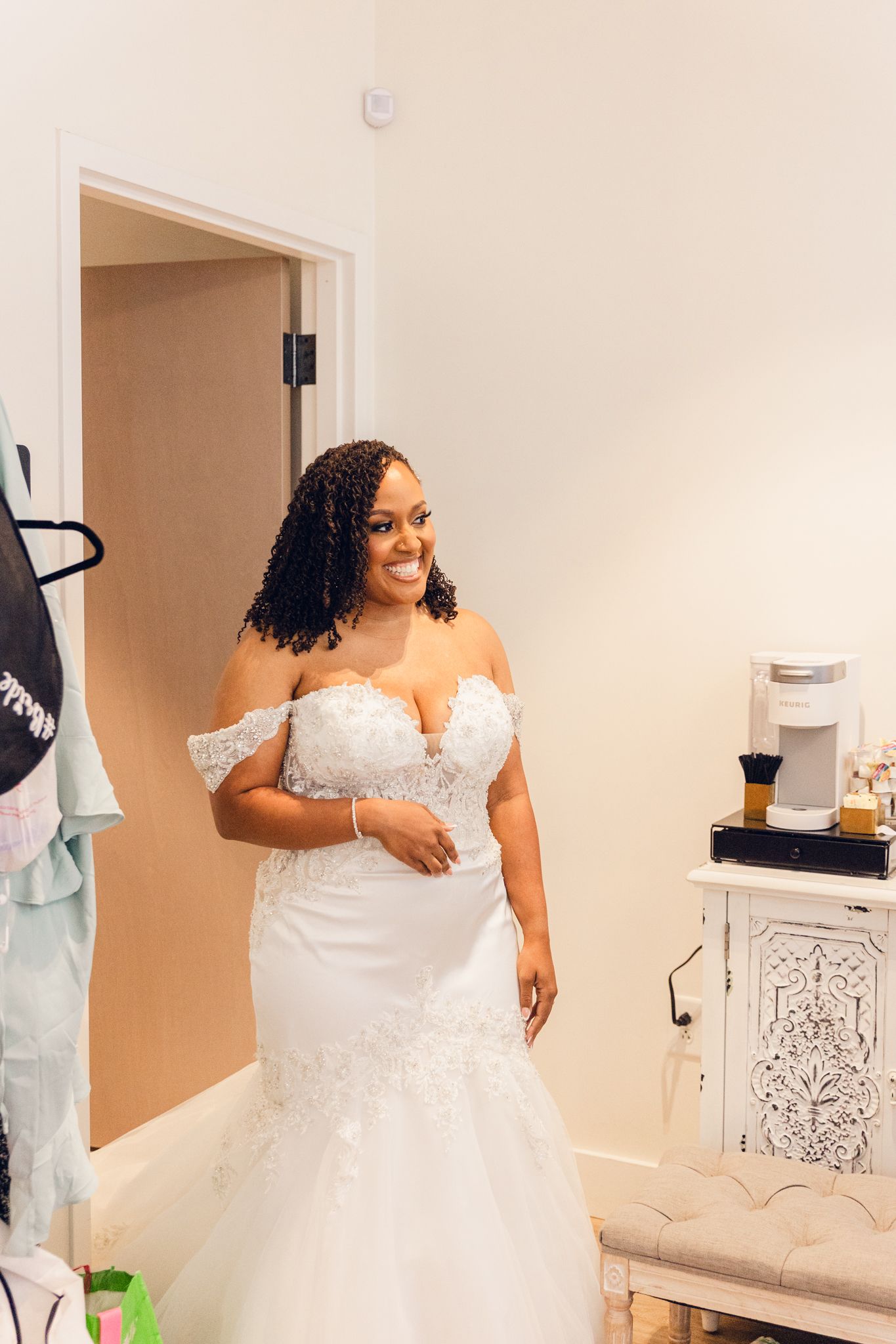 A bride in an off-shoulder white wedding gown smiles while getting ready in a dressing room.