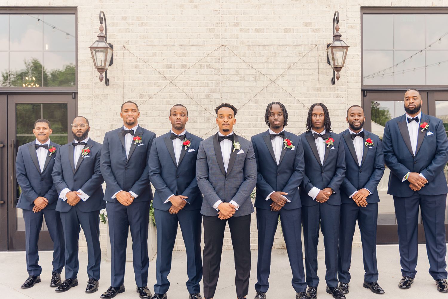 Groomsmen in navy blue suits lined up against a light colored wall at a formal wedding event.