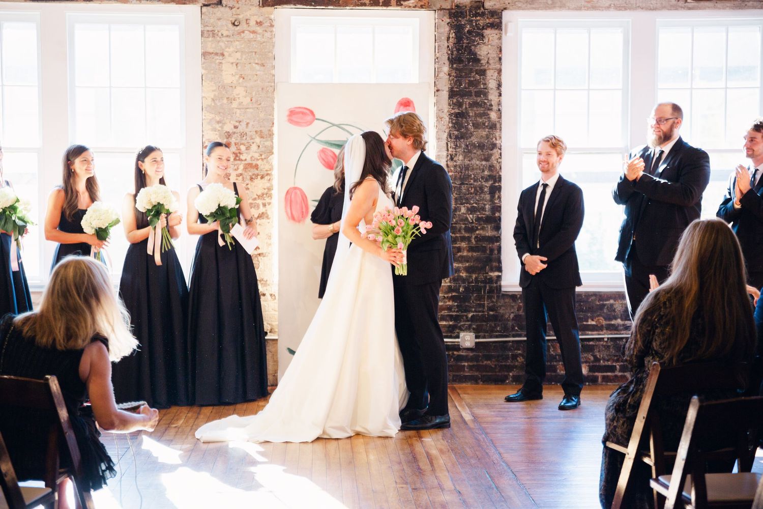 A wedding party gathers in an industrial-style venue with exposed brick walls and natural lighting.