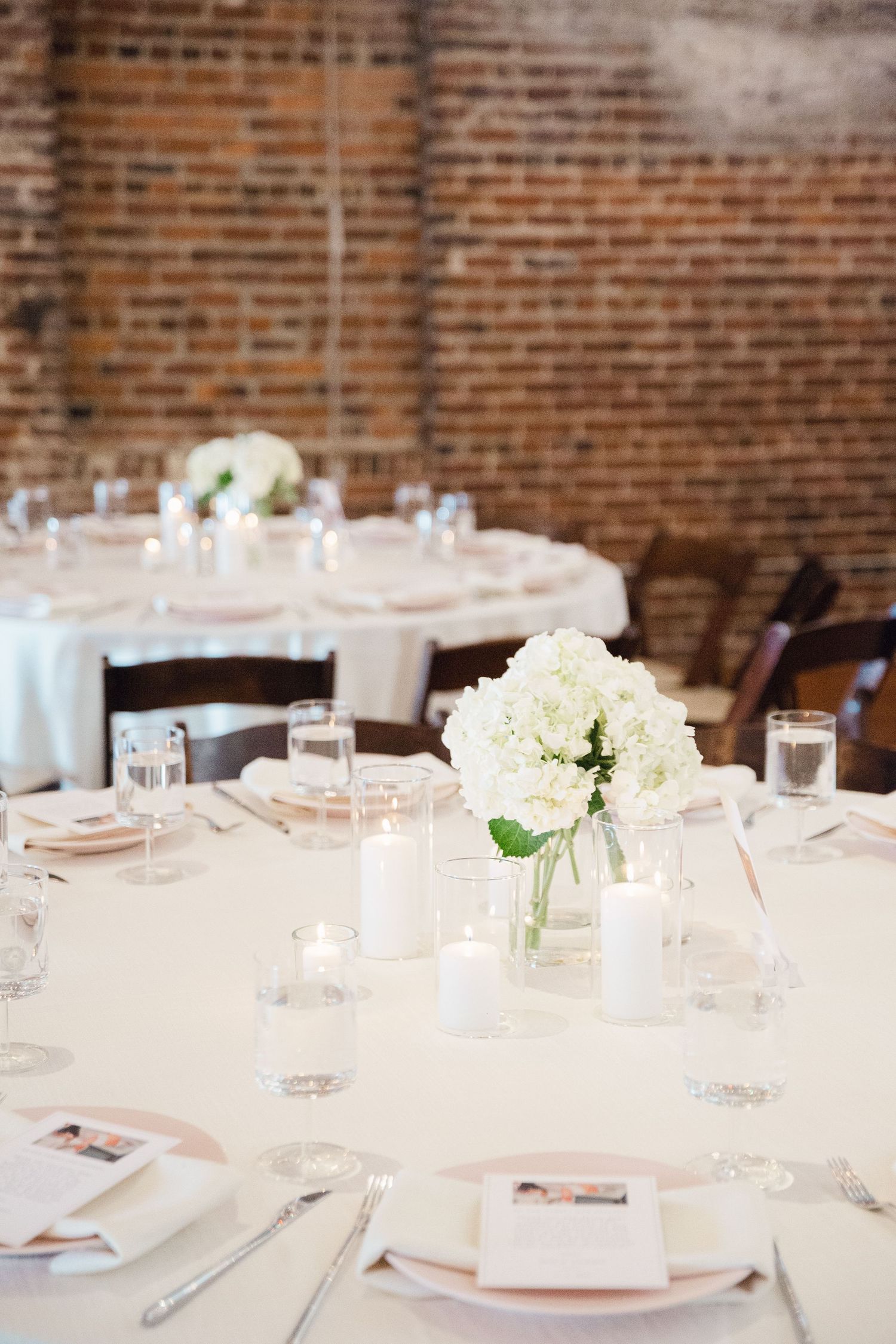 Wedding reception table with white linens, hydrangea centerpieces, and place settings against brick wall.