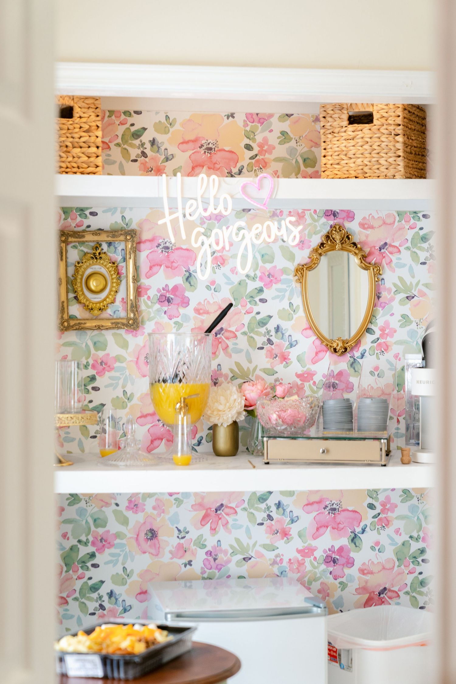 A romantic vanity area with floral wallpaper, gold mirrors, woven basket storage, and 'Hello Gorgeous' neon signage.