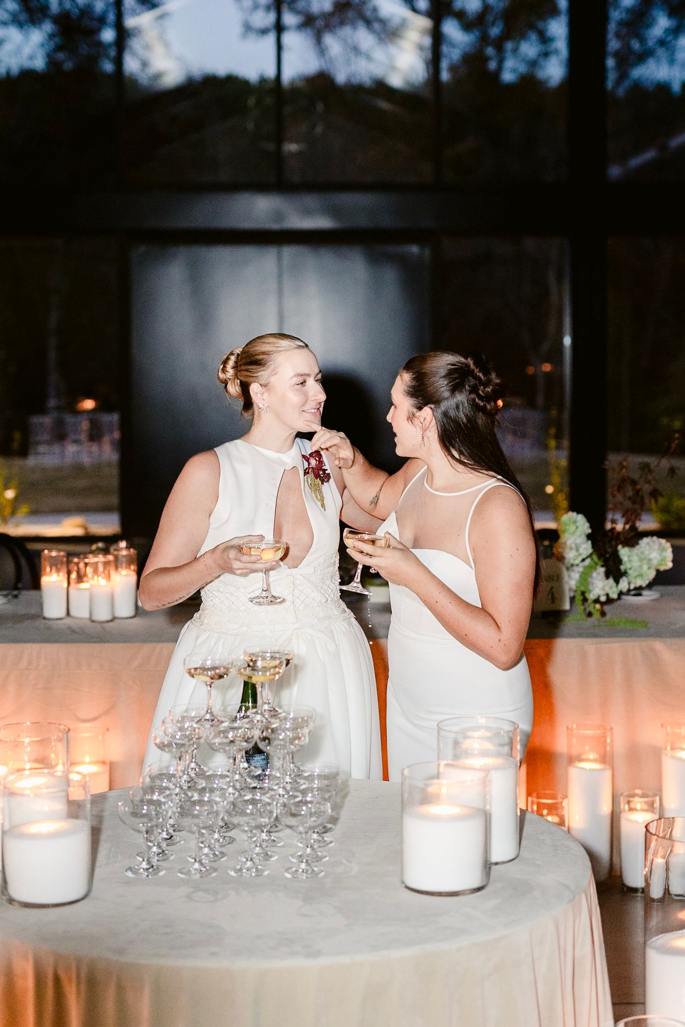 Two people in white dresses share an intimate moment at a wedding reception with glowing candles and decor.