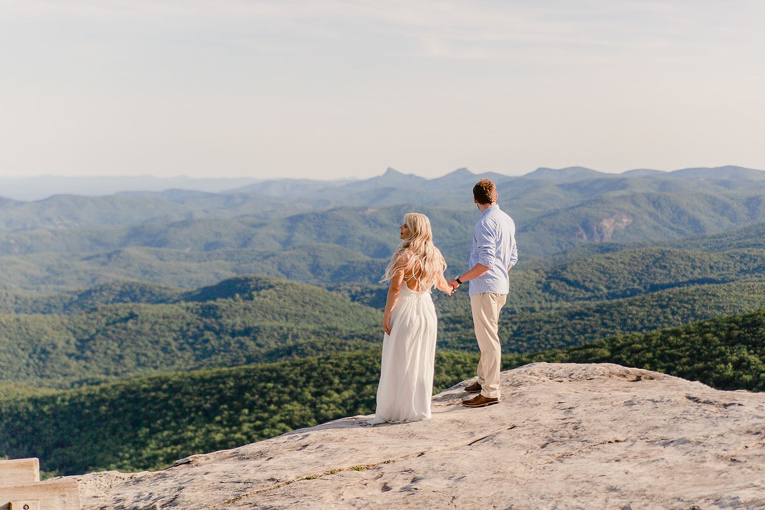 Two people stand together on rocky mountain peak overlooking vast forested landscape.