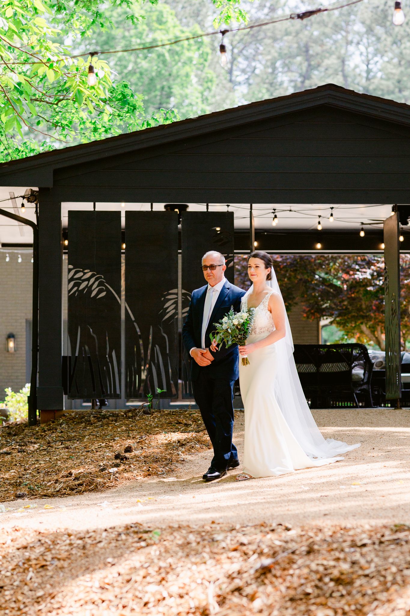 A couple in wedding attire dances together under a rustic outdoor pavilion with trees visible in the background.
