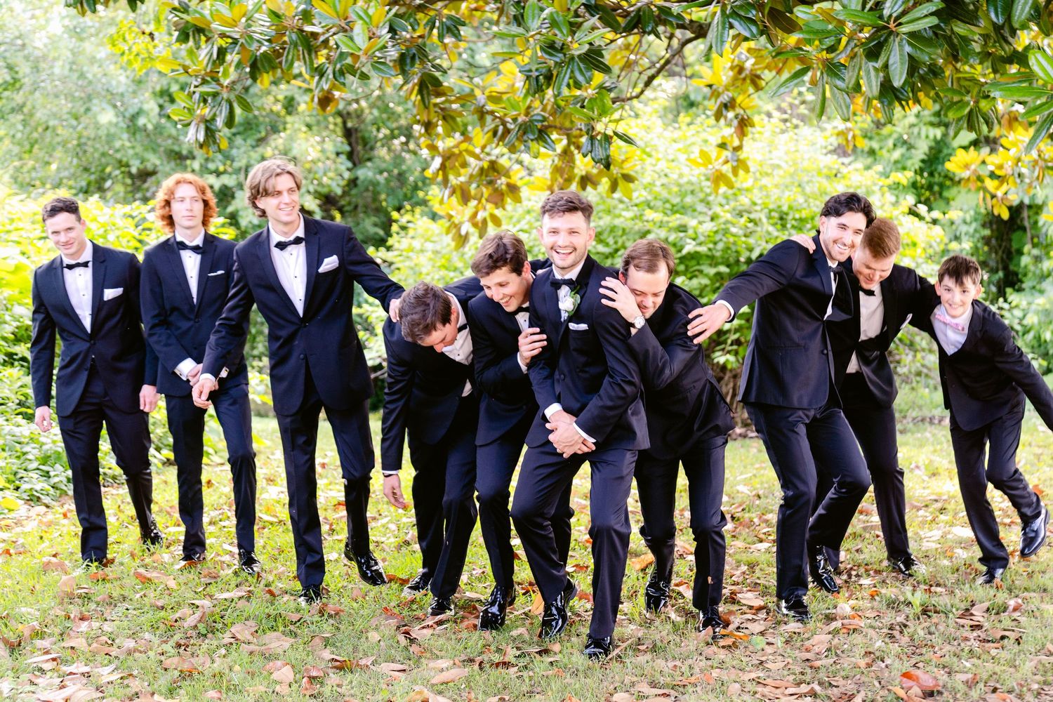 A fun wedding party in dark suits poses playfully for a creative photo series in an outdoor garden setting.