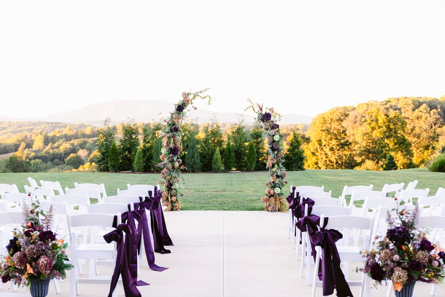 Wedding party members in purple dresses and suits walk in pairs down an outdoor aisle with white chairs.