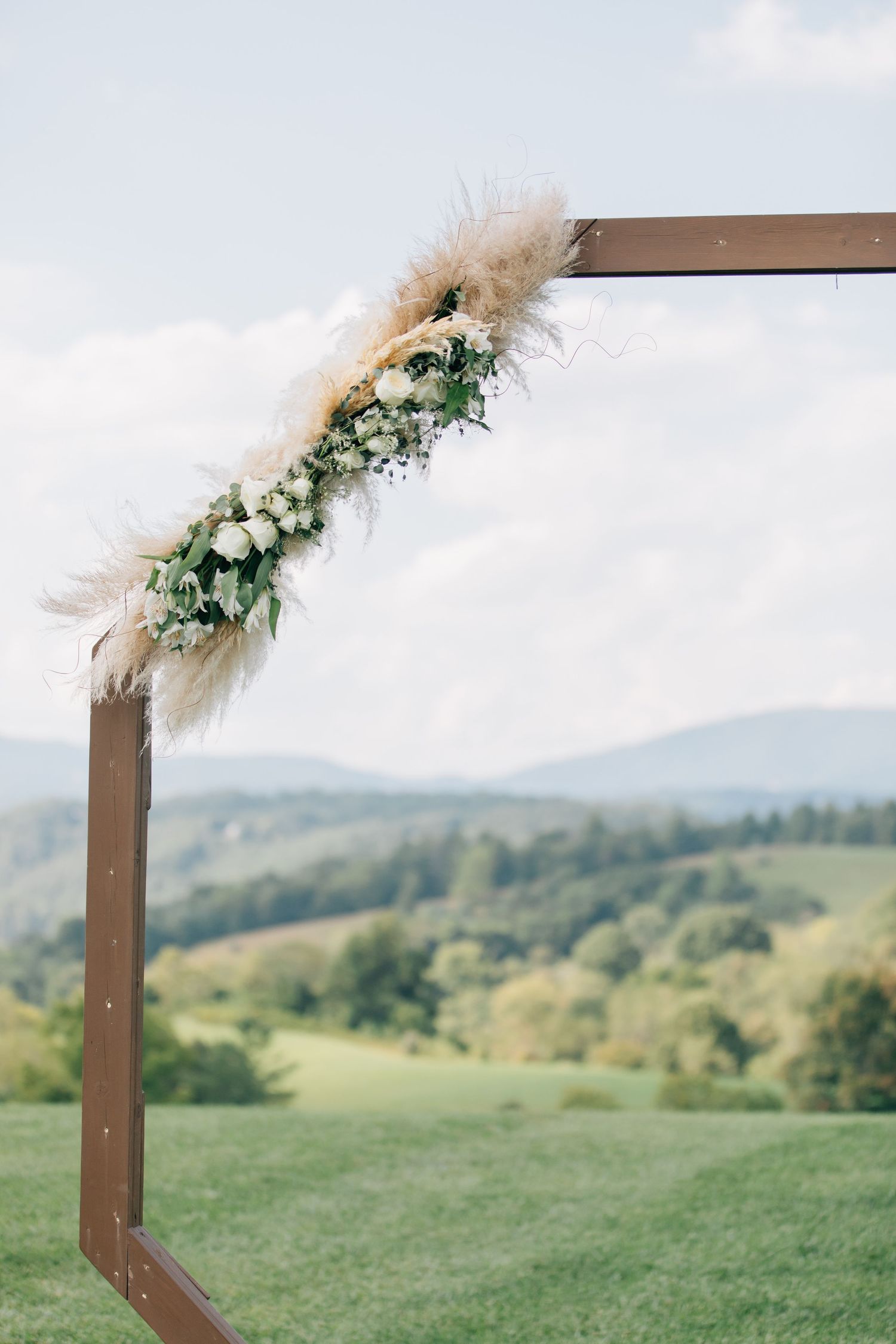 Wooden wedding arch decorated with pampas grass and white flowers stands on a green hilltop with mountains in background.